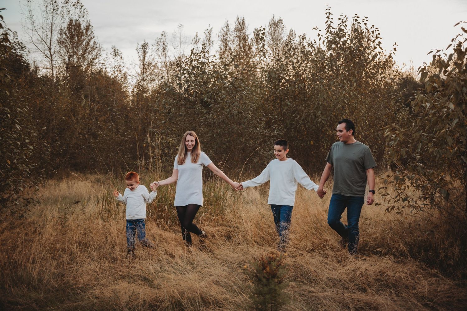 Happy family of four holding hands walking through tall golden grass in autumn nature.