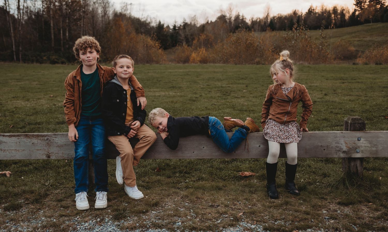 Four children sit and play on a wooden fence rail in an open autumn field.