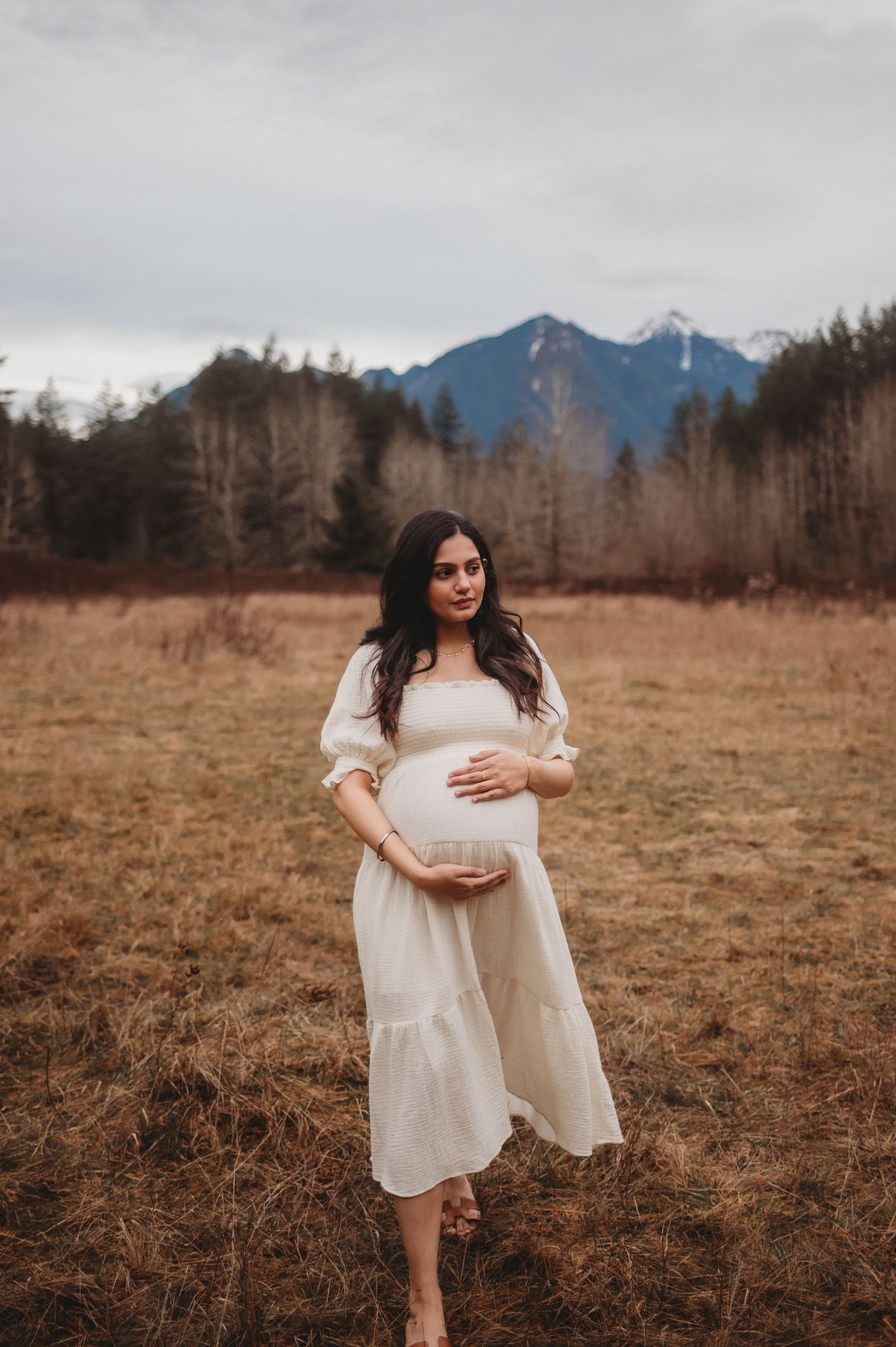Pregnant woman in cream dress cradles baby bump in golden field with mountain backdrop.