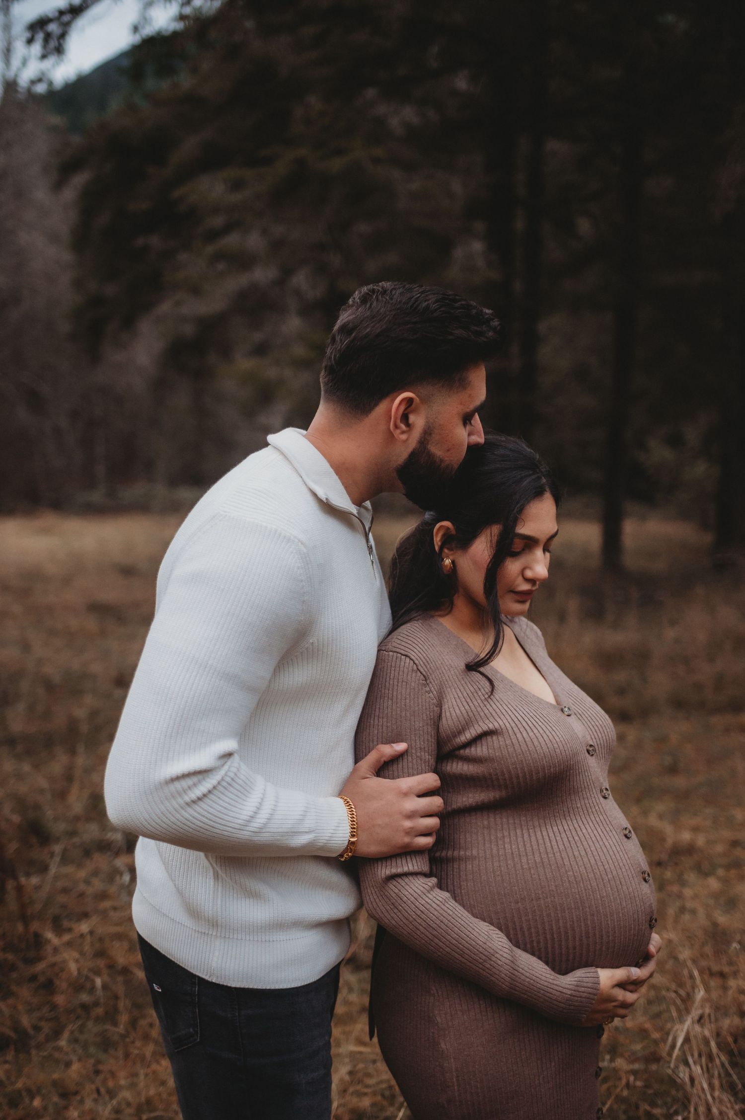 Man in white sweater embracing pregnant partner from behind in rustic outdoor fall maternity shoot.