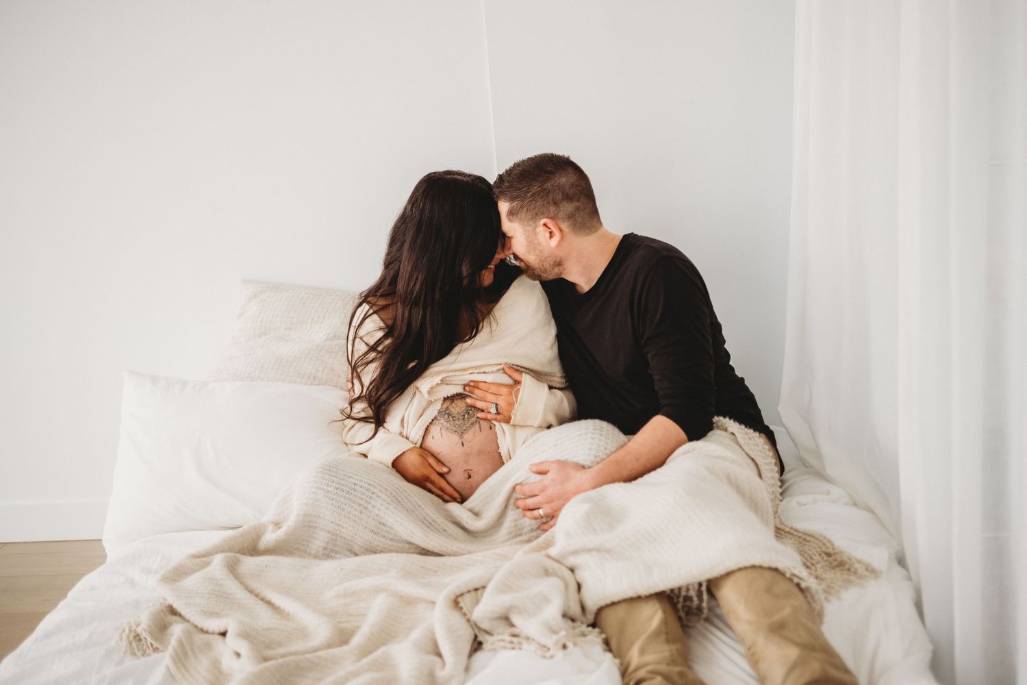 Couple kissing on white bed with pregnant belly during maternity session.