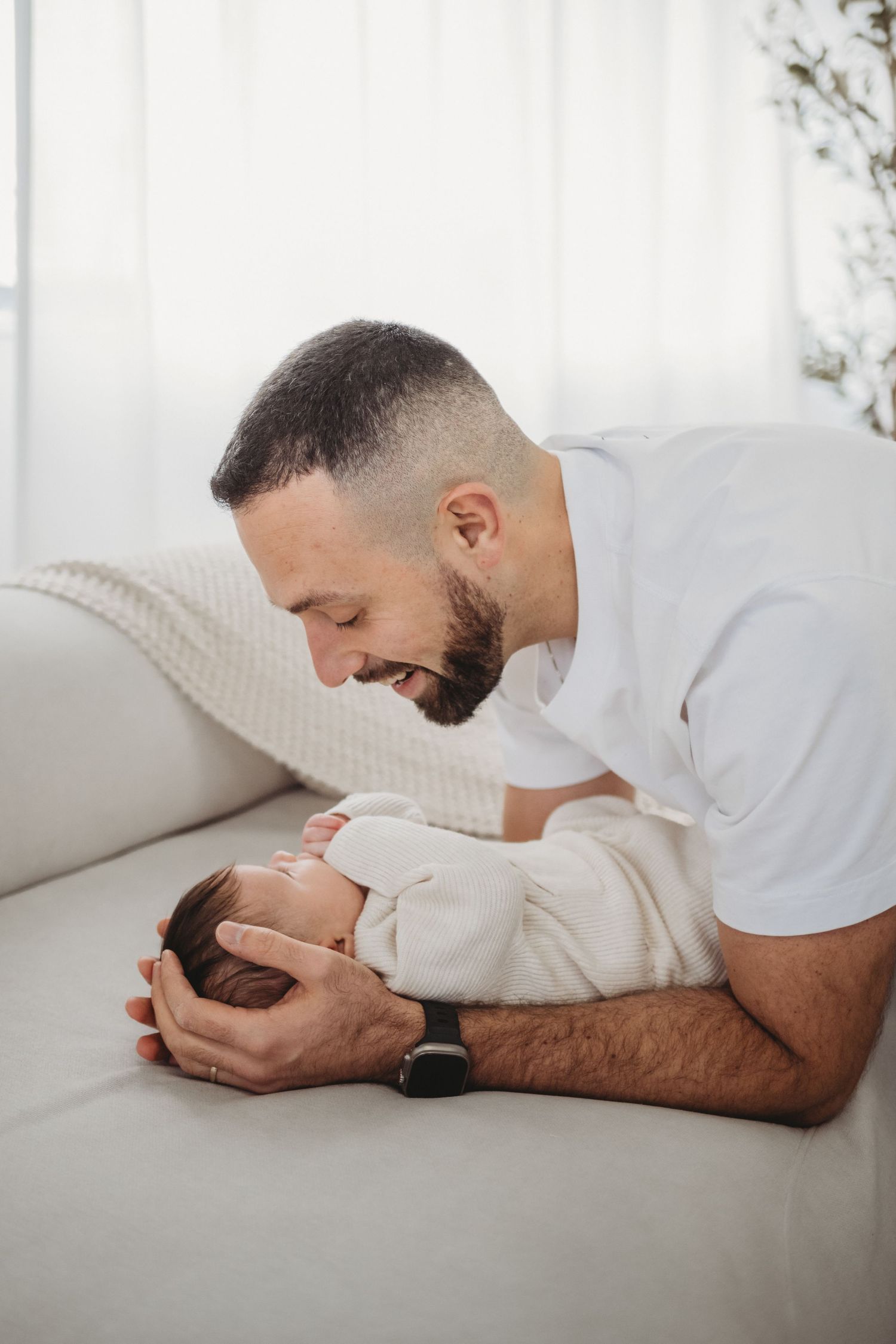 Bearded father smiling and bonding with newborn baby on white sofa.