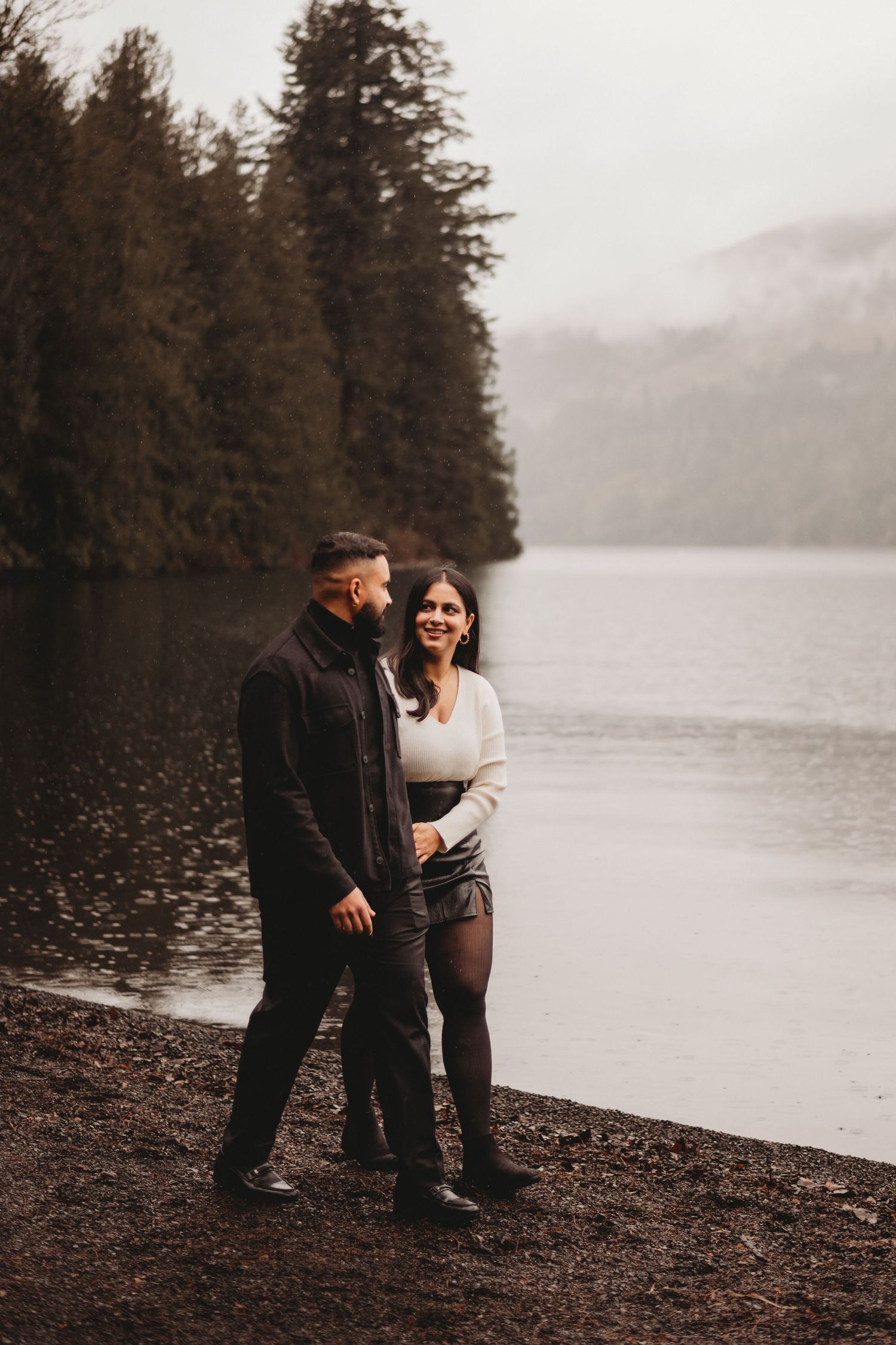 Couple standing by a misty lake surrounded by evergreen trees during an overcast day.