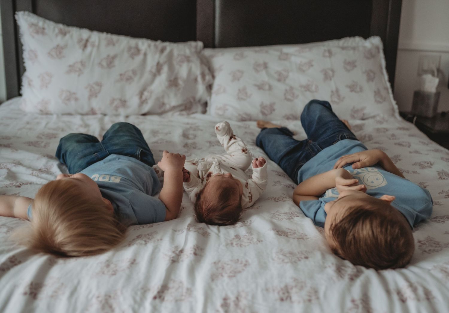 Three young children lying on a bed with floral bedding, playing together in matching blue outfits.