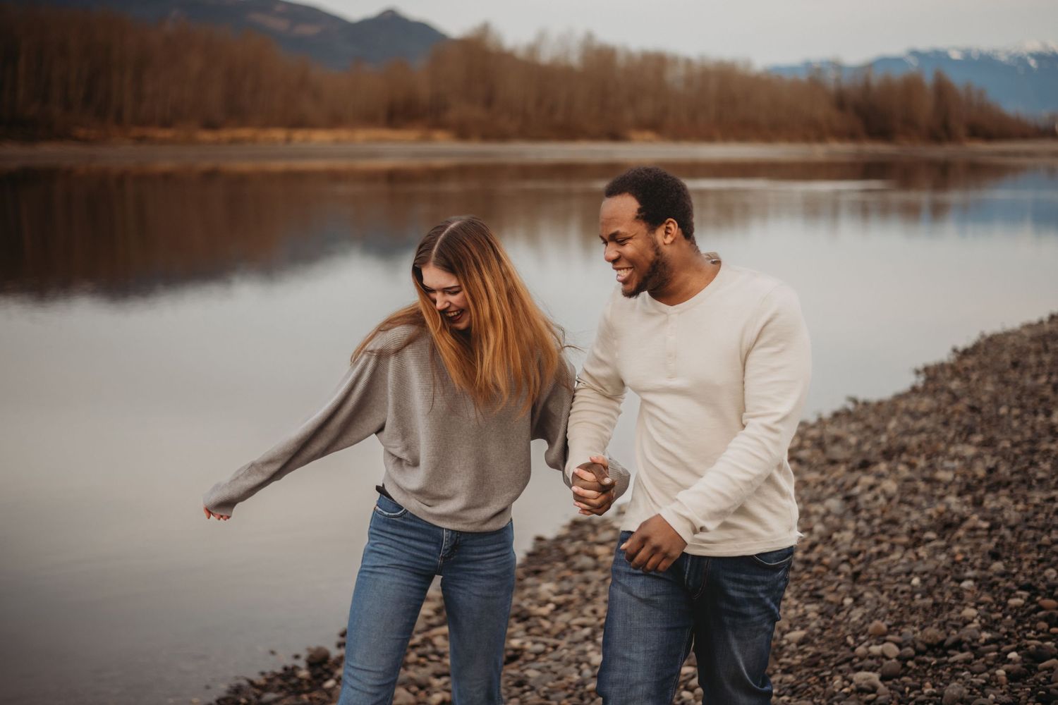 Laughing interracial couple holding hands and walking along a scenic rocky riverbank with mountain views.