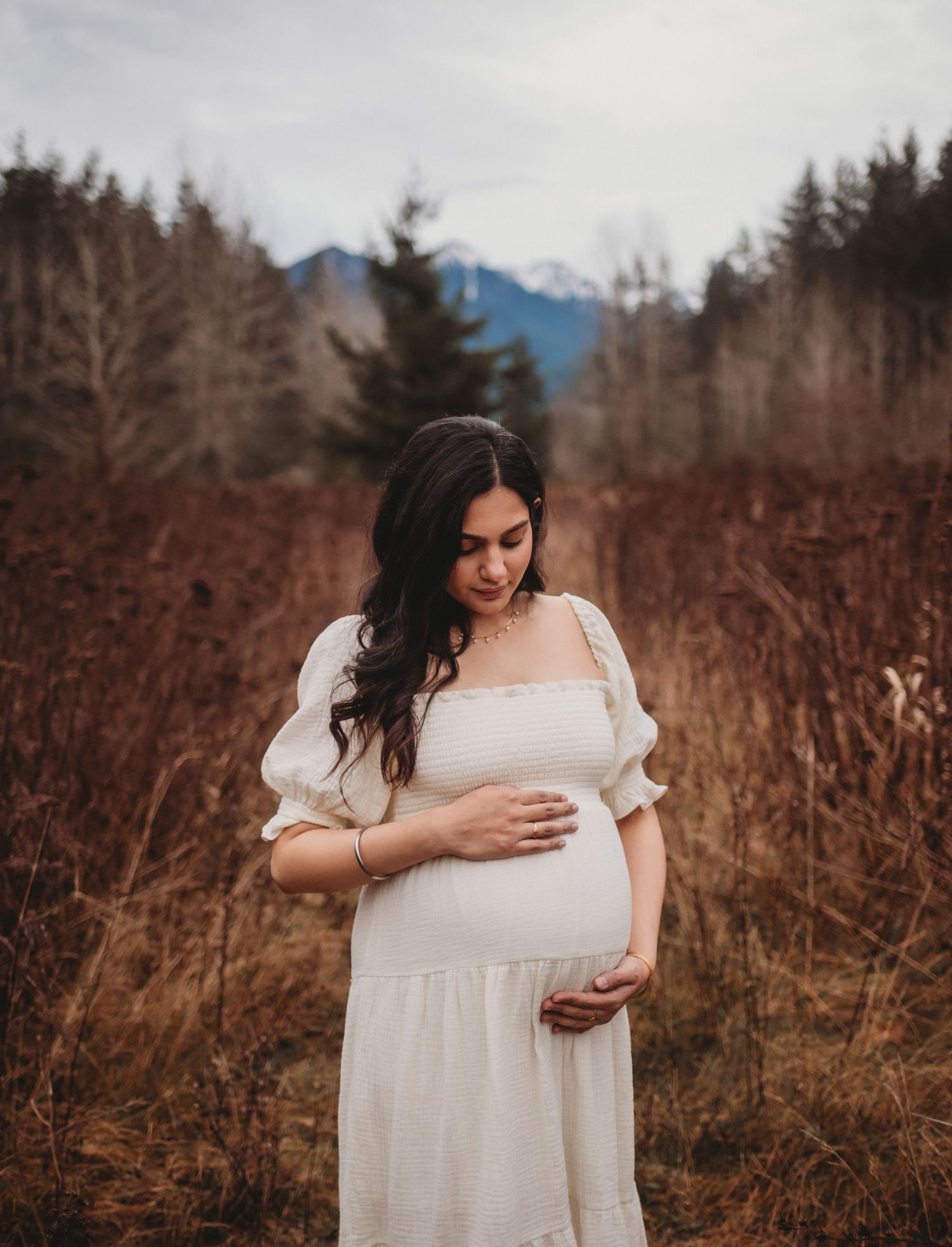 Pregnant woman in white puff-sleeve dress cradles bump among autumn grasses with mountain view.