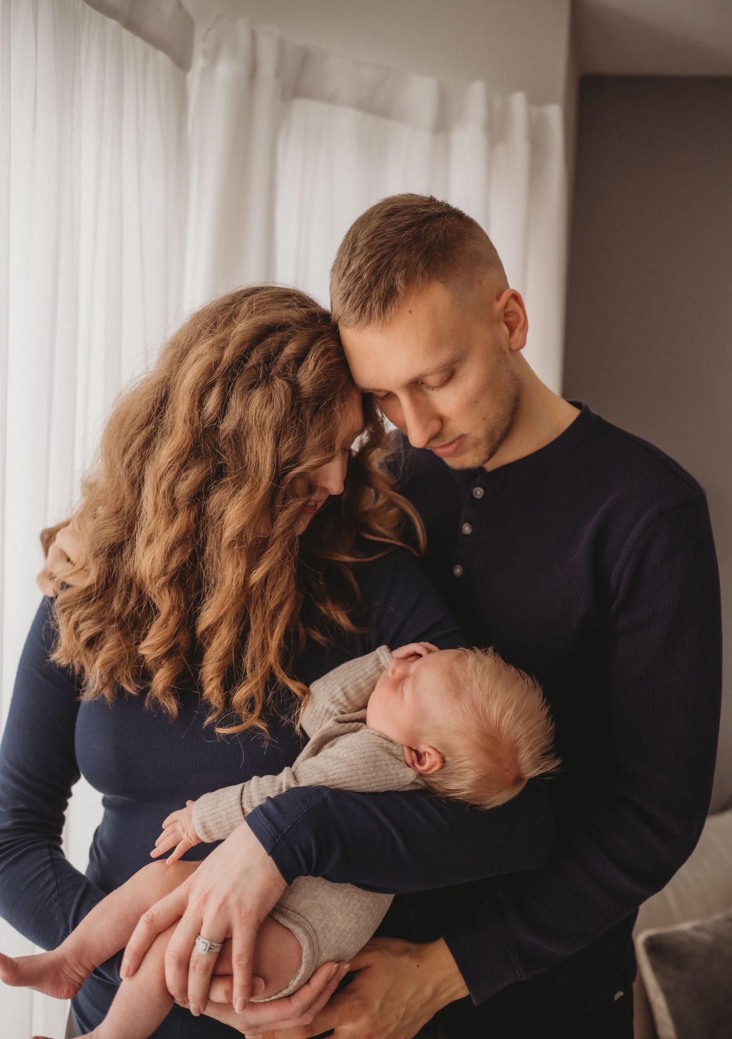 Young couple touching foreheads lovingly while holding their sleeping newborn baby together by window.