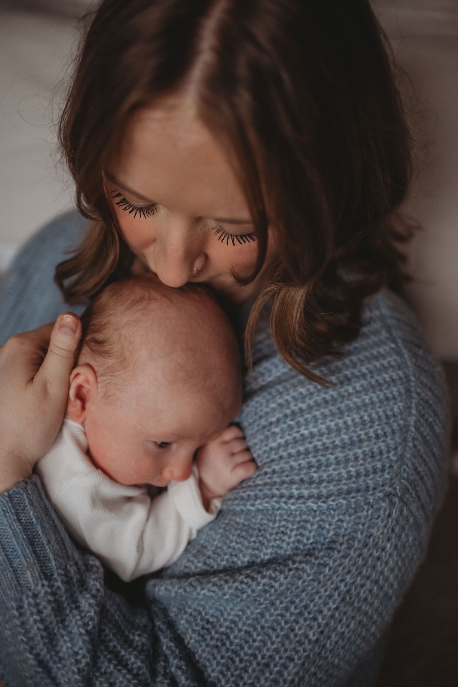 Mother tenderly kissing her sleeping newborn baby wrapped in a blue knit blanket.