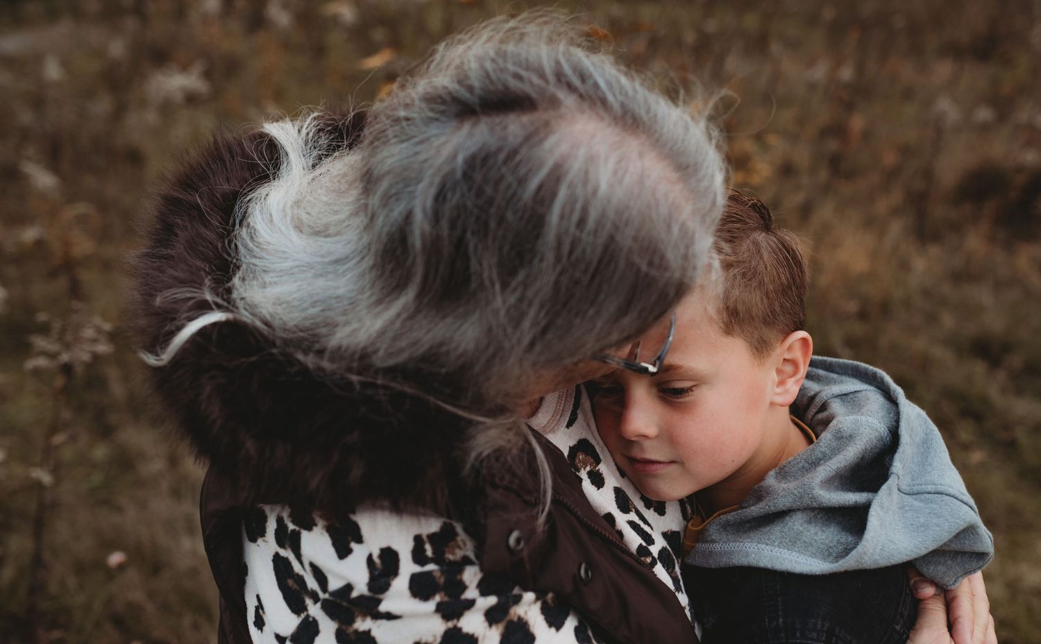 A woman with gray hair tenderly embraces a young boy outdoors in a field during autumn.