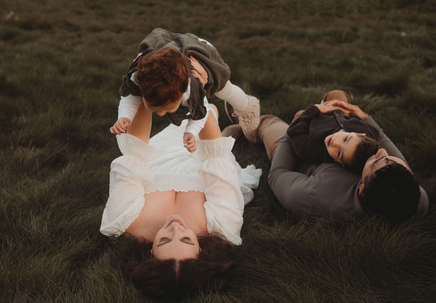 A family of four lies on green grass, parents playing with their two young children in a candid moment.