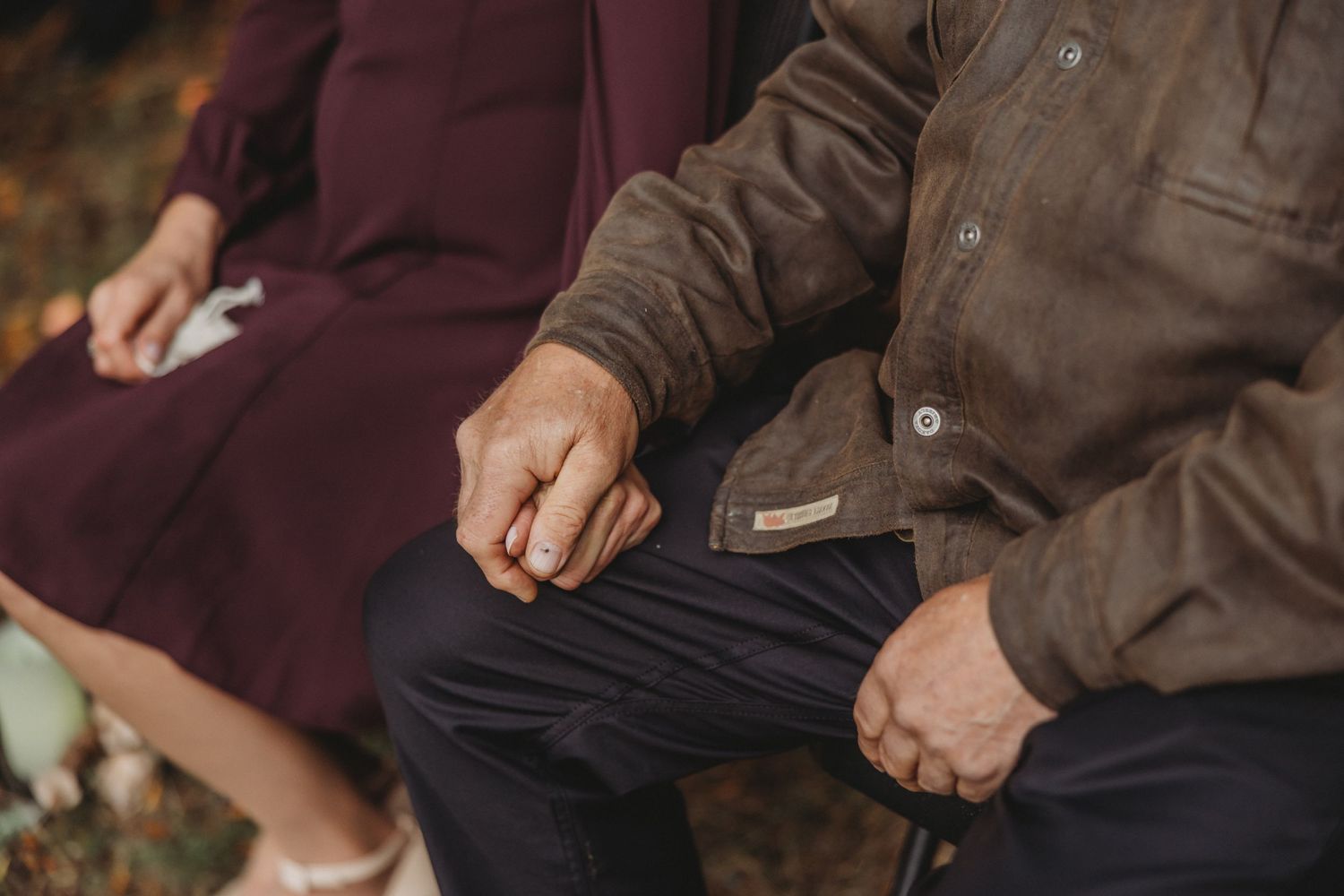 Close-up of elderly couple holding hands tenderly while seated outdoors in autumn setting.