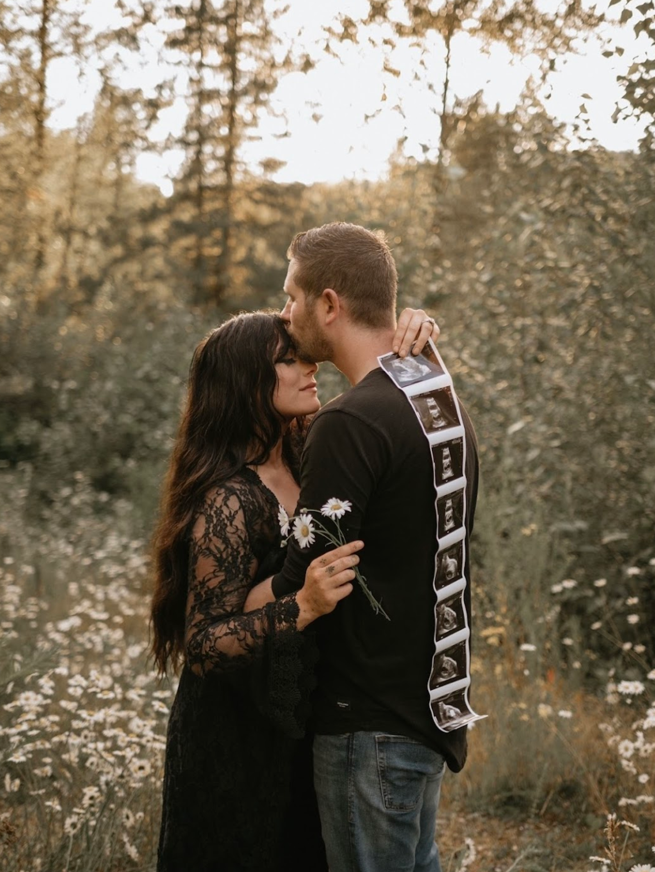 A couple shares a tender kiss outdoors while the man holds ultrasound photos, announcing their pregnancy in nature.