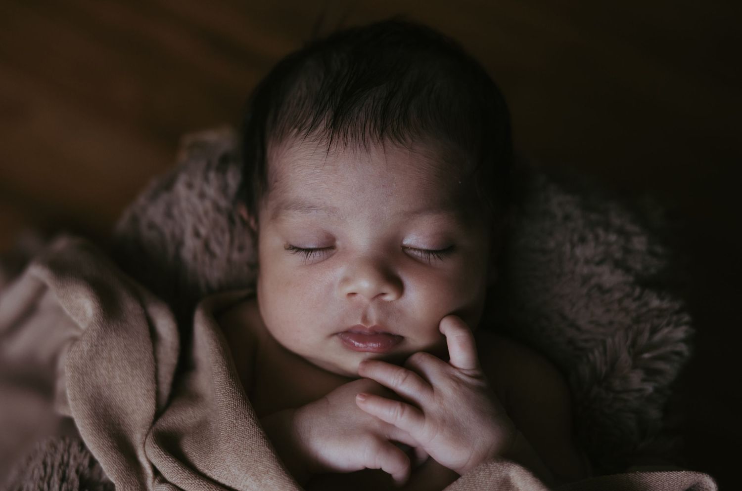 Sleeping newborn baby nestled in a cozy brown knit wrap with hands tucked under chin.
