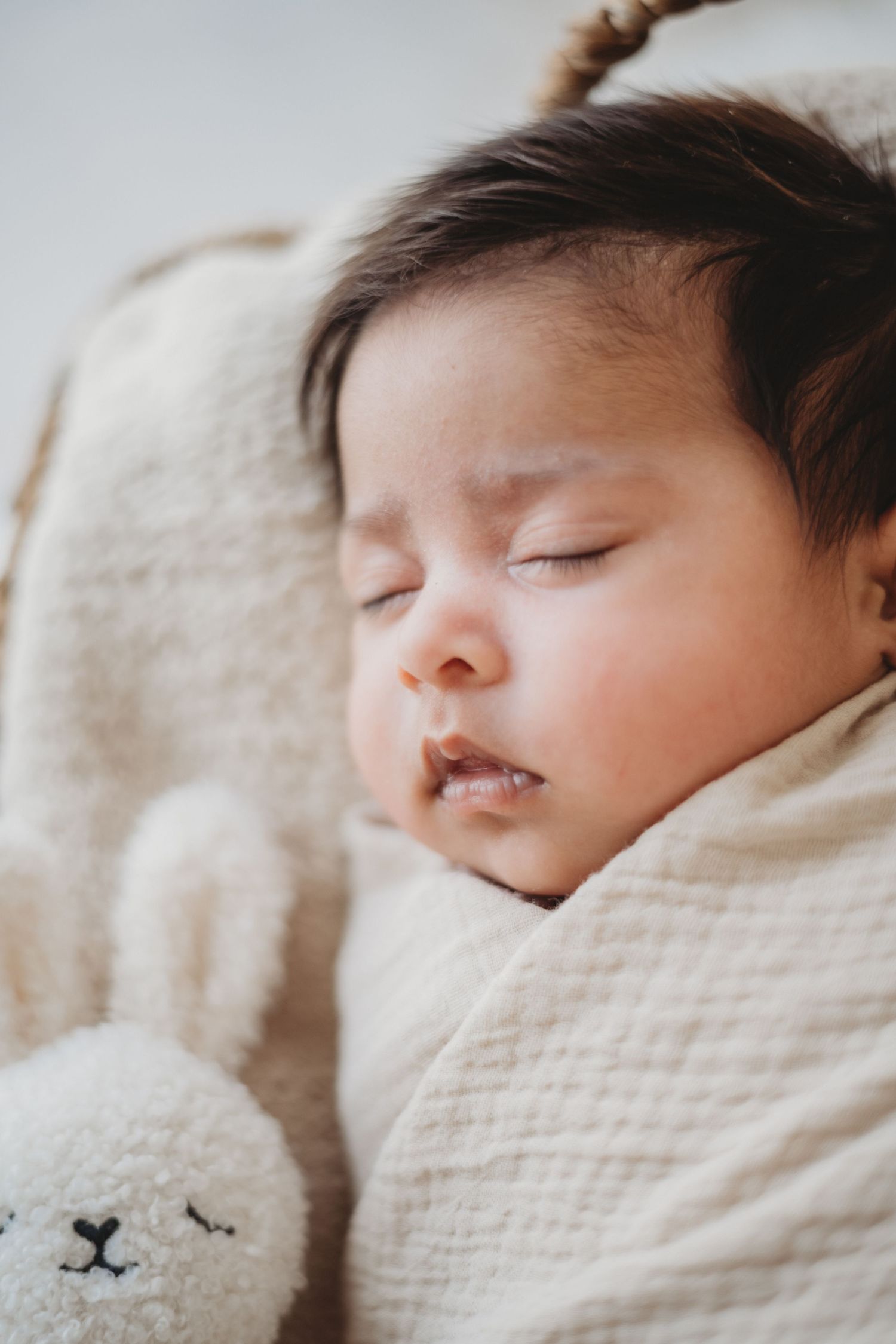 Peaceful sleeping newborn baby swaddled in cream wrap next to soft bunny stuffed toy.