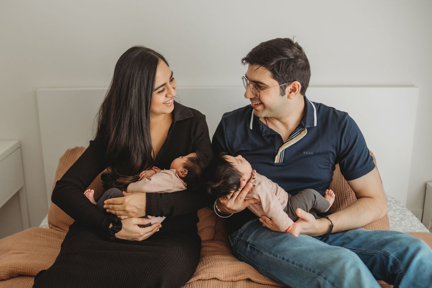 Smiling parents sitting on a bed each holding a newborn twin baby, looking at each other lovingly.