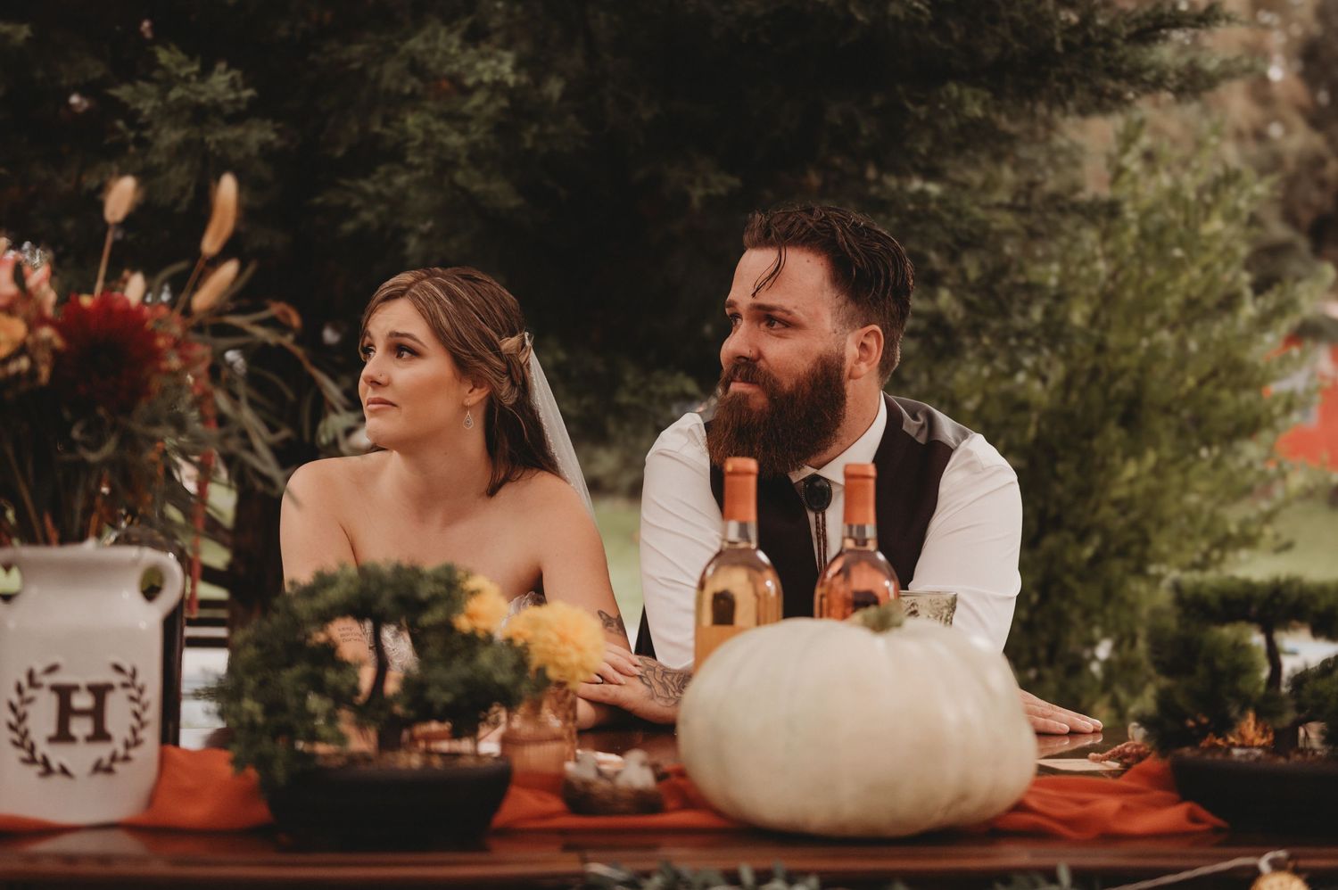 A bride and bearded groom sit together at a fall-themed wedding table with a white pumpkin centerpiece.