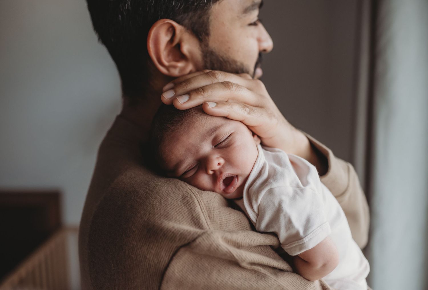 A bearded father gently holds his sleeping newborn baby against his shoulder.