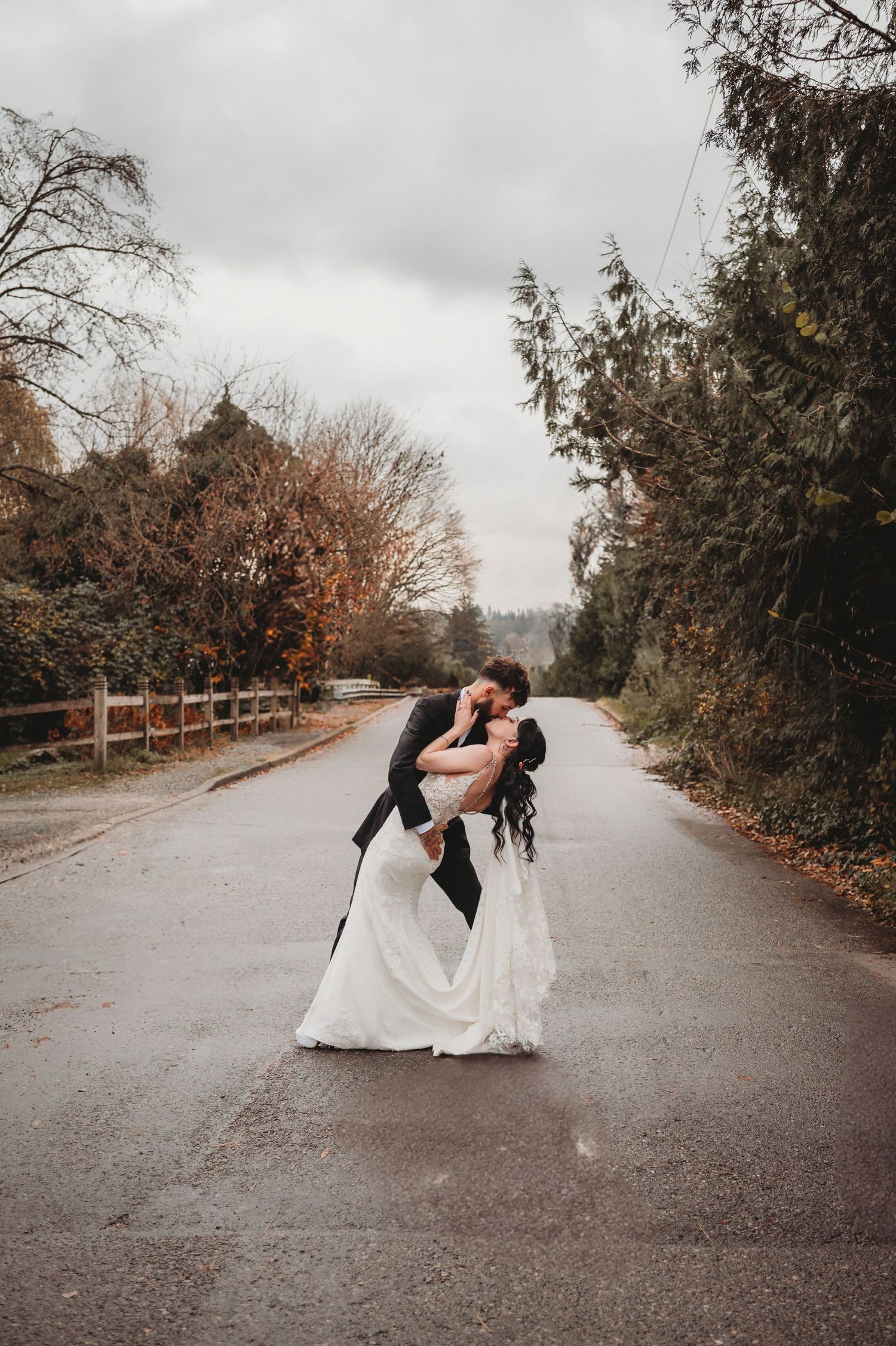 Bride and groom share a romantic dip kiss on a rain-soaked road surrounded by autumn trees.