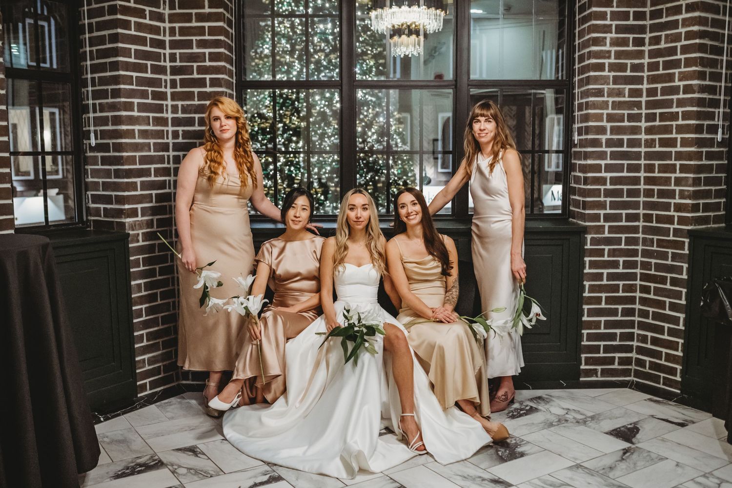 Bride poses with four bridesmaids in champagne dresses inside a chic brick-walled venue.