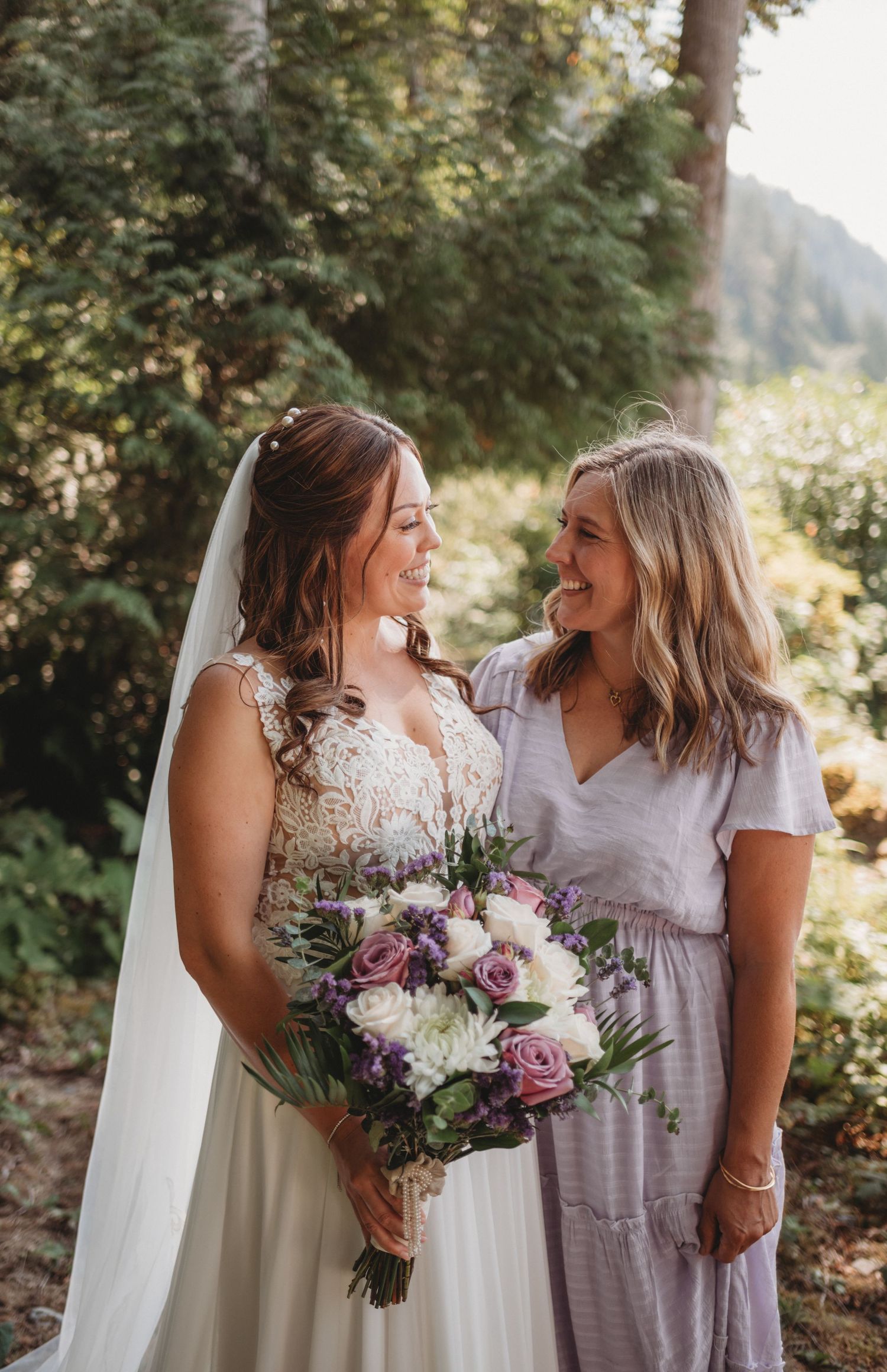 Smiling bride in lace gown holds purple bouquet with female companion.