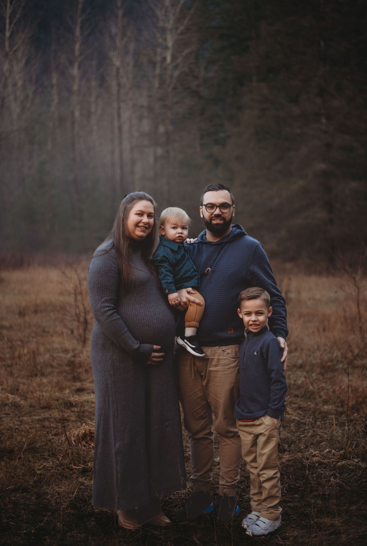 Family portrait with pregnant mother, father holding toddler, and young son in forest field.