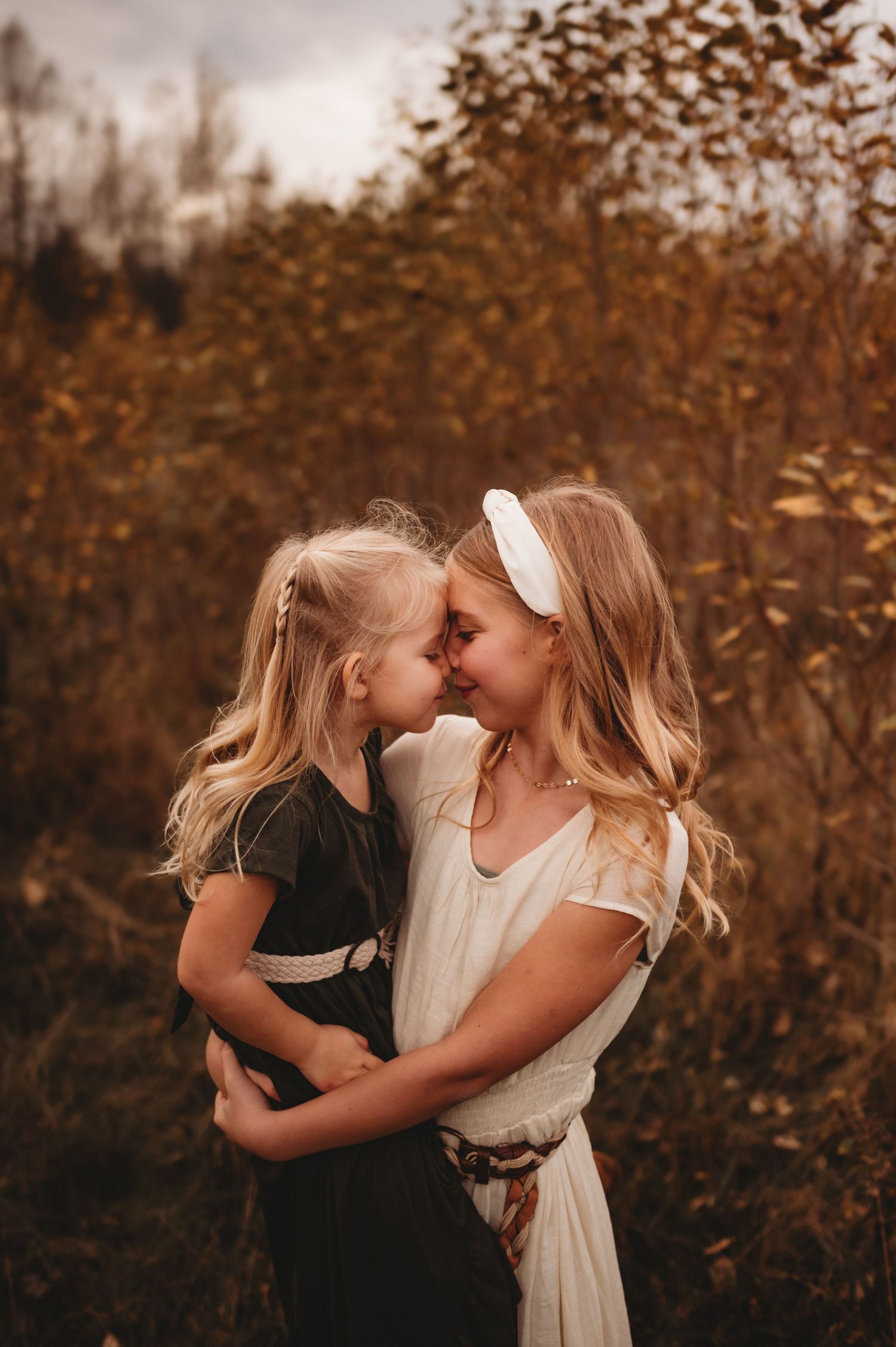 Blonde mother and daughter touching foreheads in tender embrace among autumn foliage outdoors.