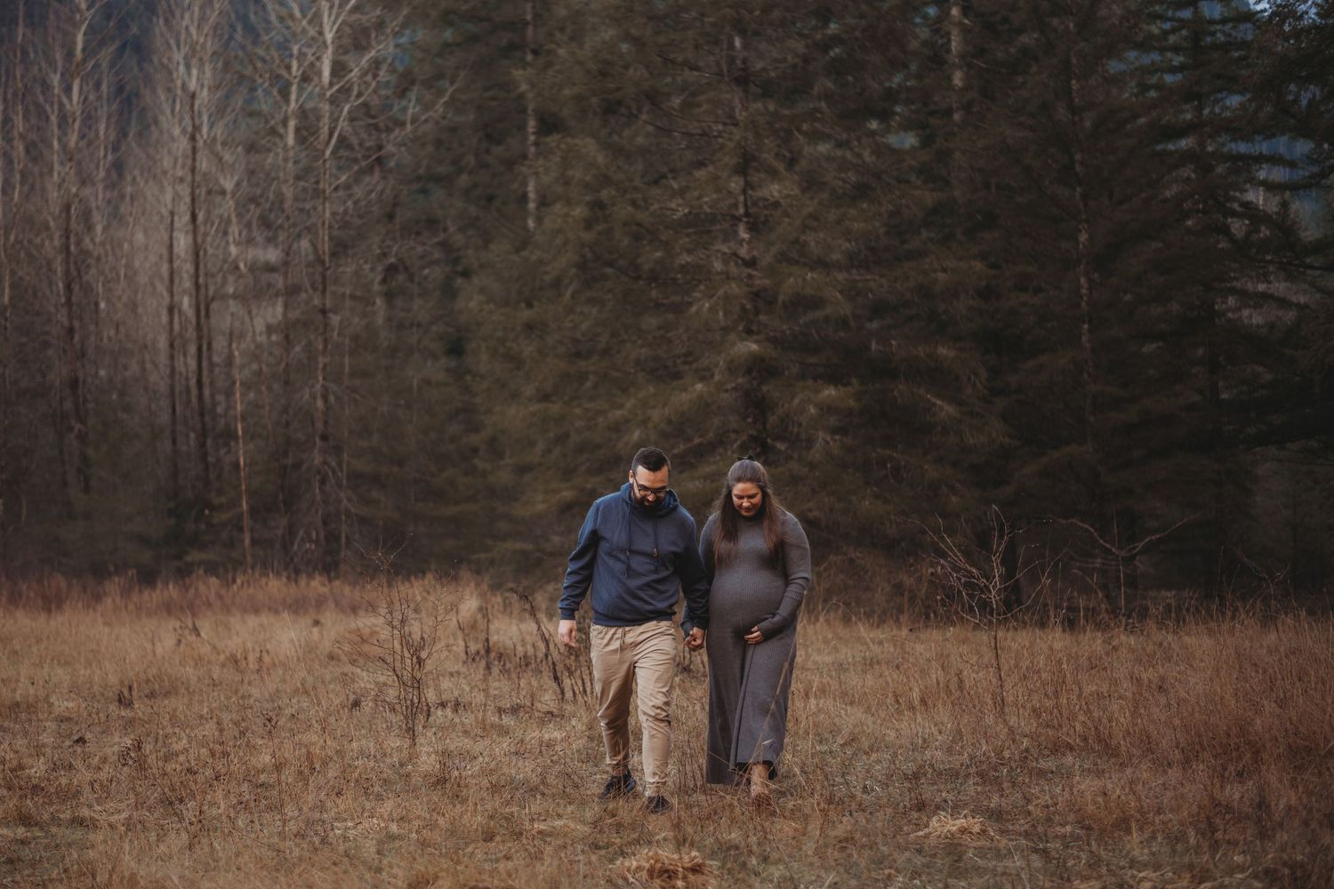 Couple walking hand in hand through dry grass field with forest and mountains in background.