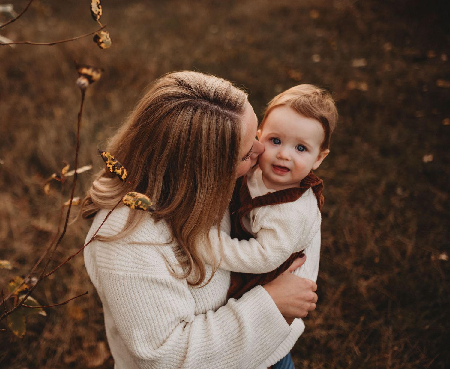 A mother in a white sweater kisses her toddler on the cheek in a fall meadow.