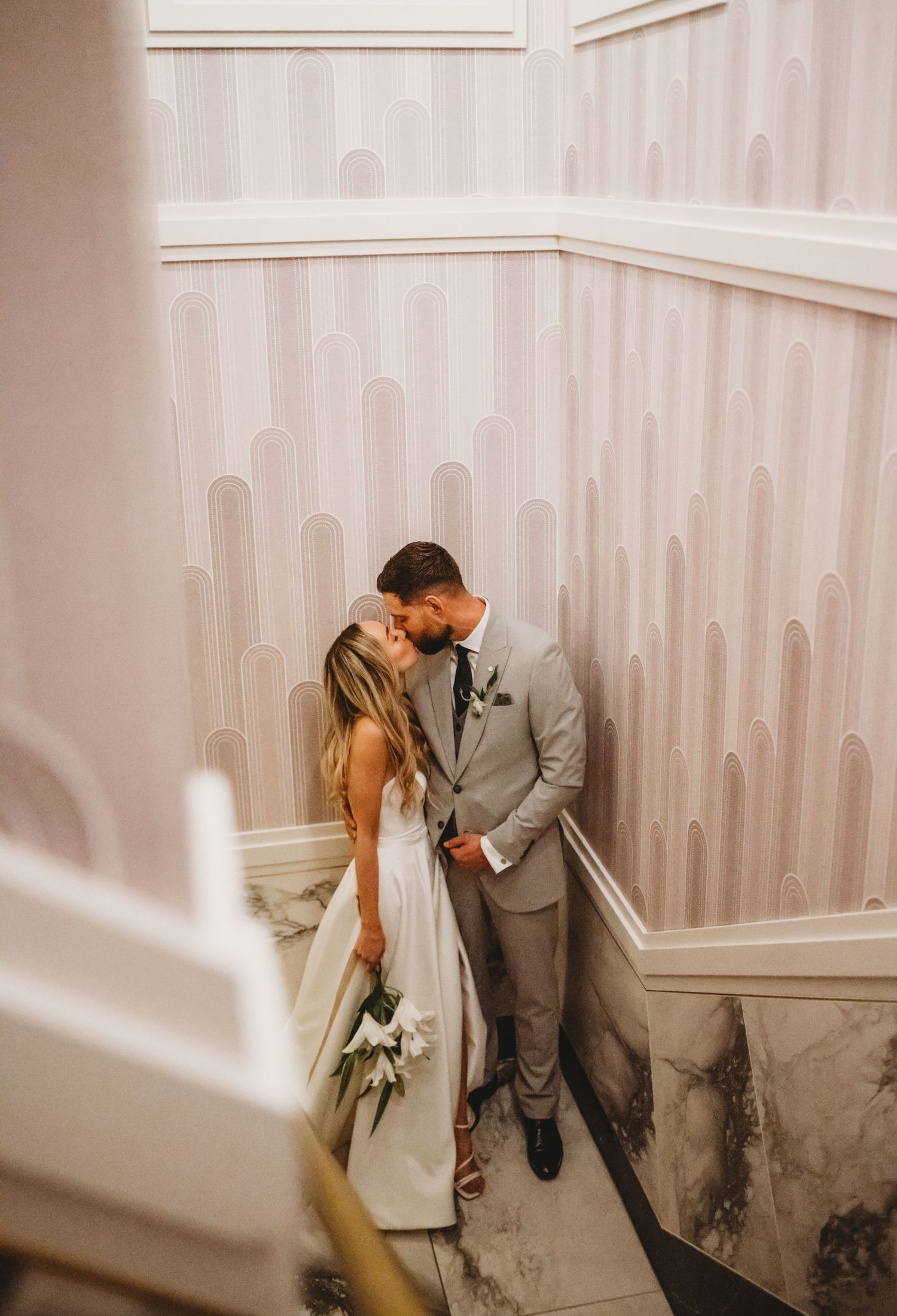 Bride and groom share a romantic kiss on a marble staircase with elegant pink wallpaper.