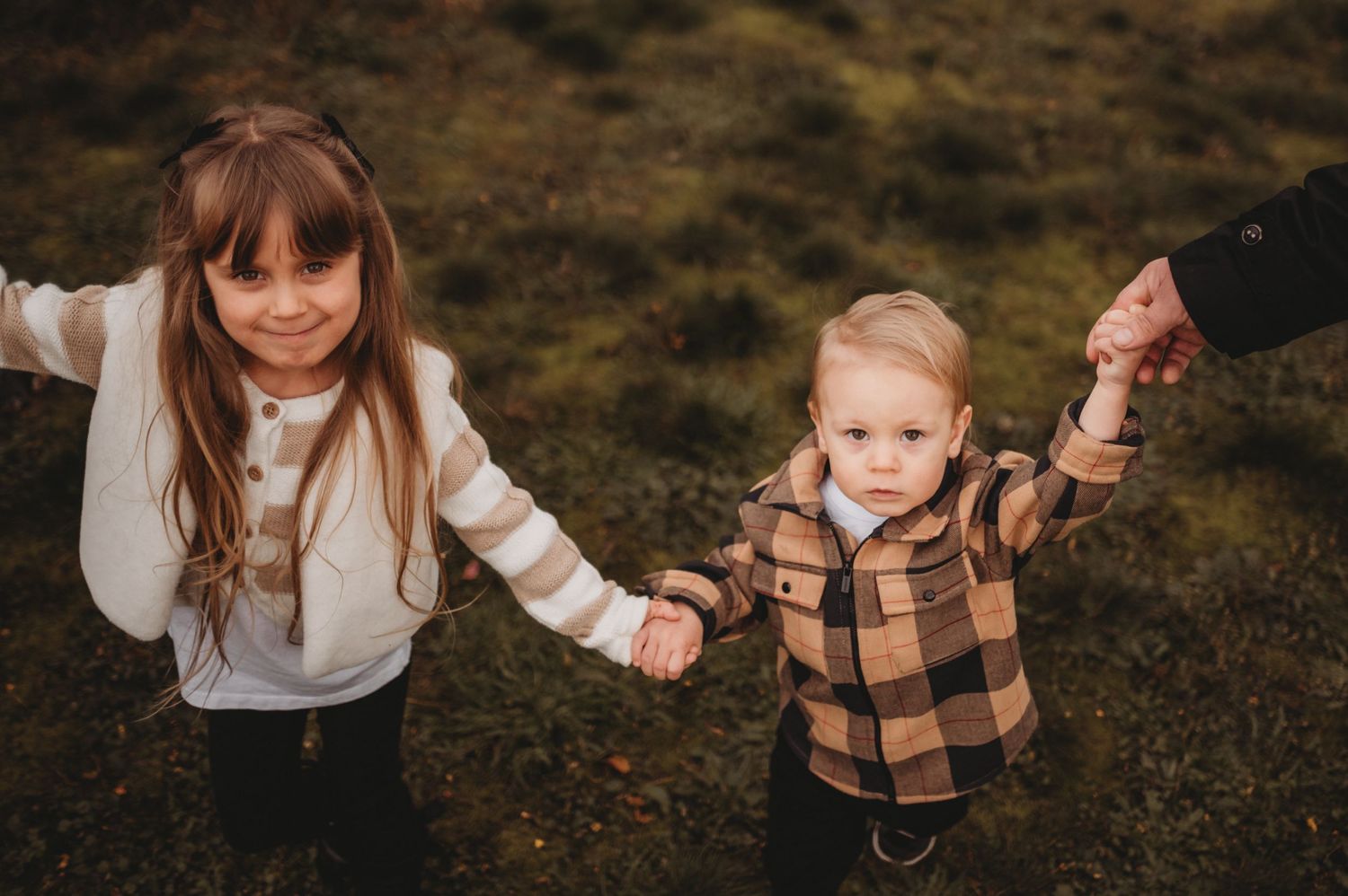 Two young siblings holding hands in green mossy outdoor setting.