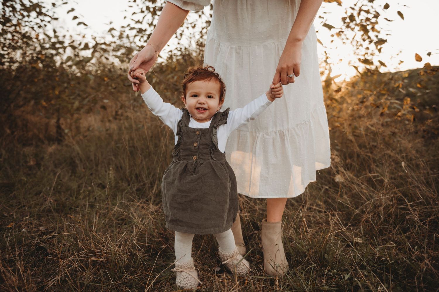 Toddler girl learning to walk held by mother outdoors.