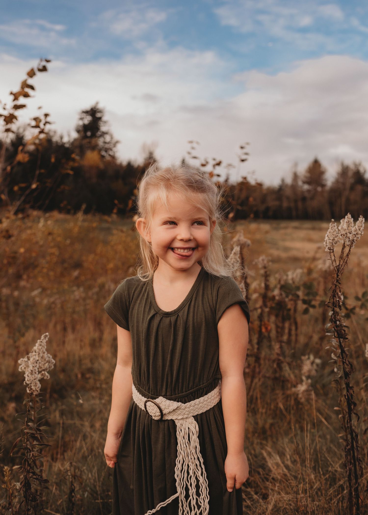 Adorable blonde girl in olive dress with macrame belt smiling in autumn field under blue sky.