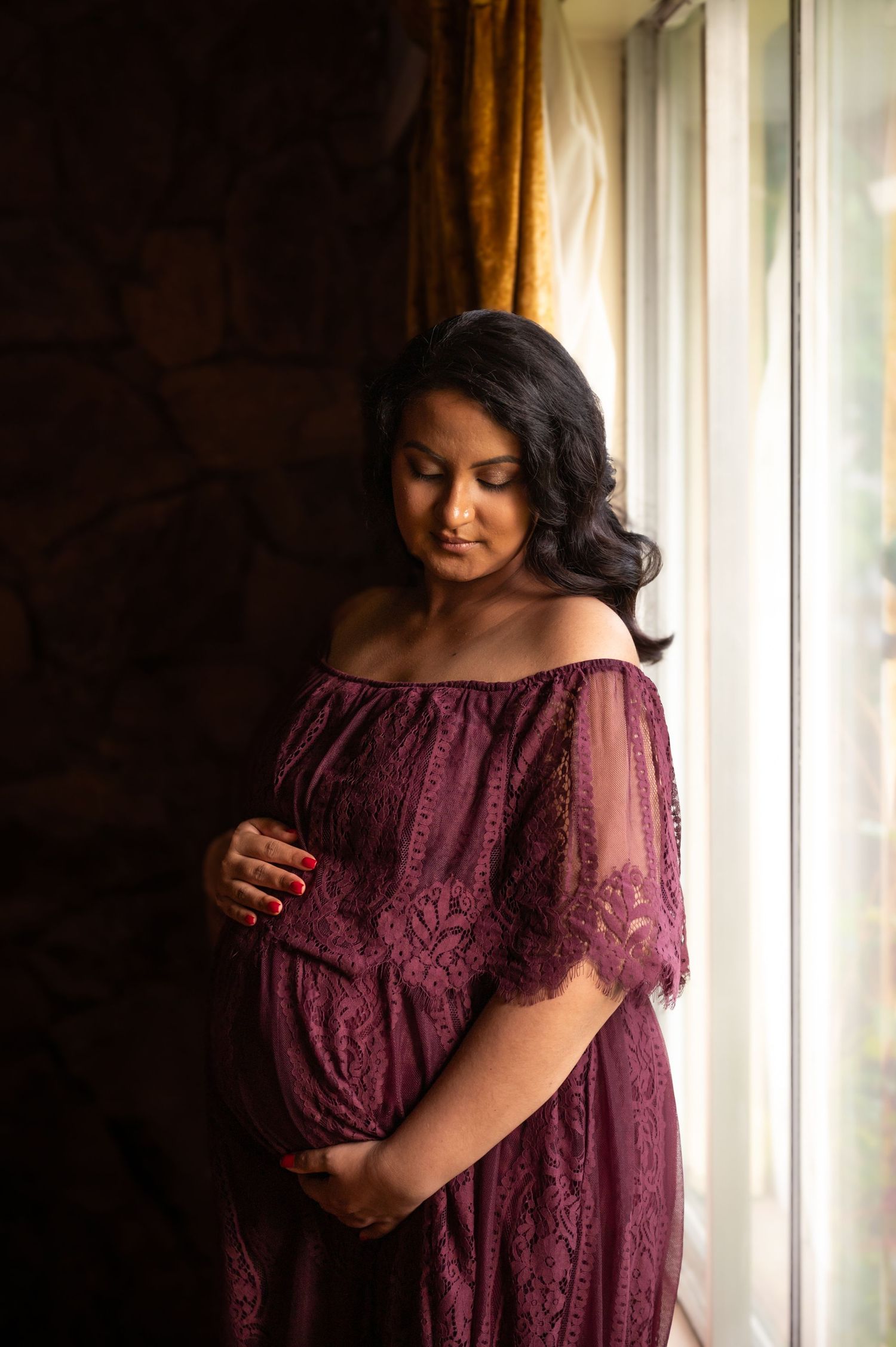 Pregnant woman in burgundy lace off-shoulder gown cradling baby bump by window.