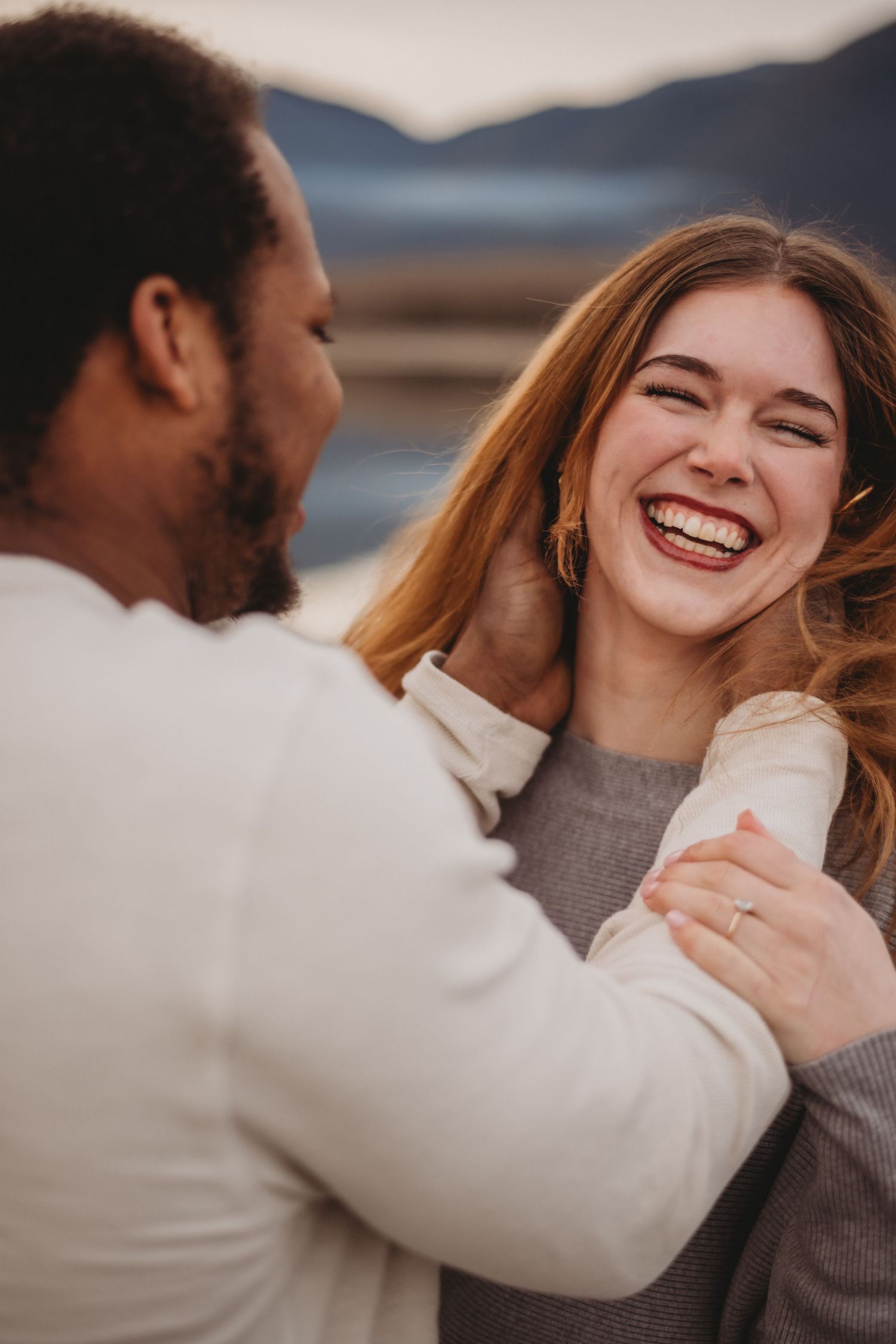 Laughing woman with engagement ring enjoys a romantic moment with her partner by a lake.