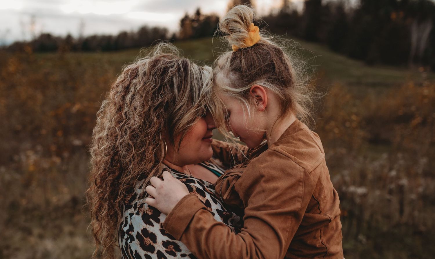 A mother and young daughter share a tender forehead-to-forehead embrace in an autumn field.