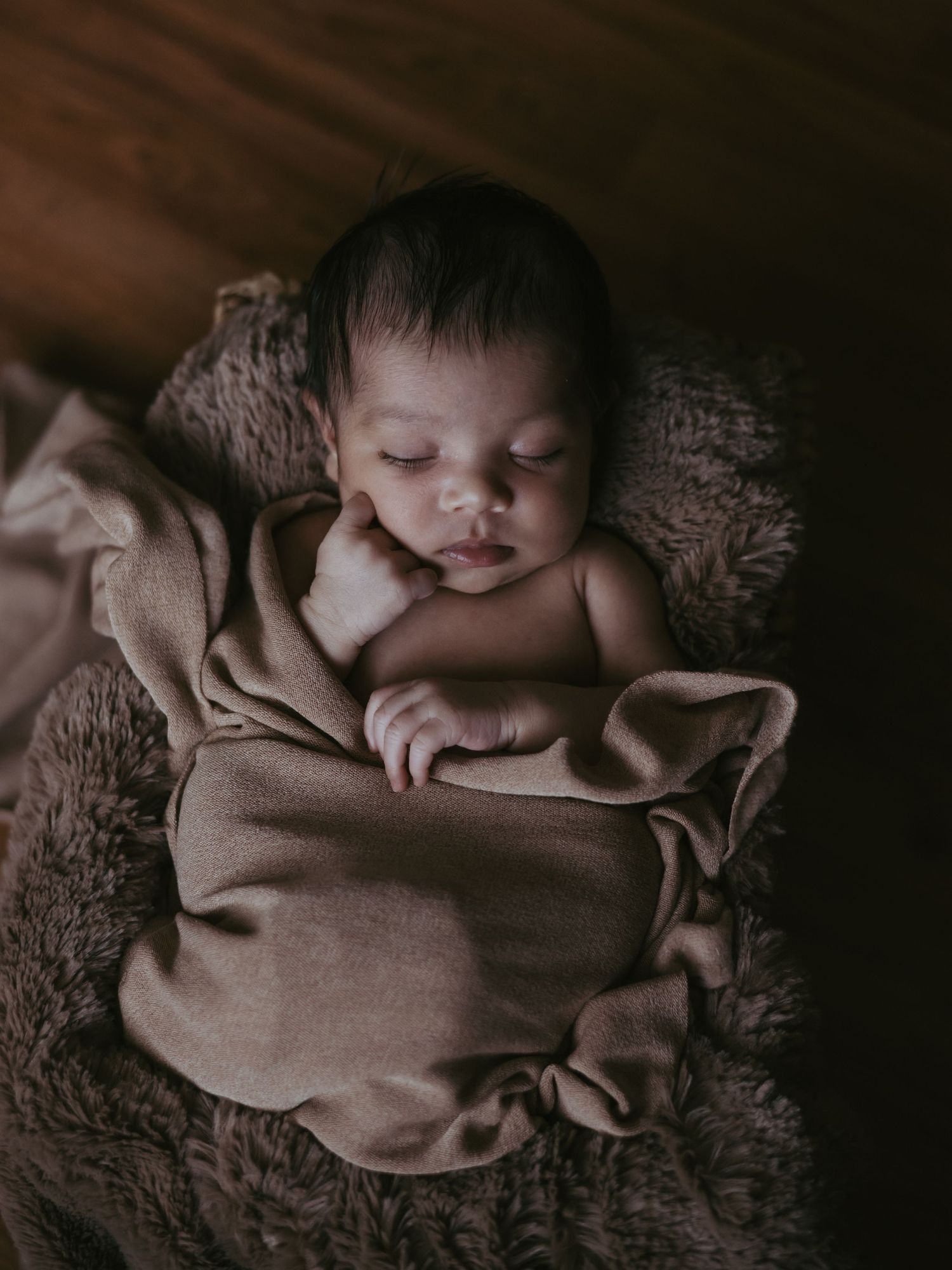 Newborn baby sleeping curled up in a brown wrap inside a fluffy basket in warm lighting.