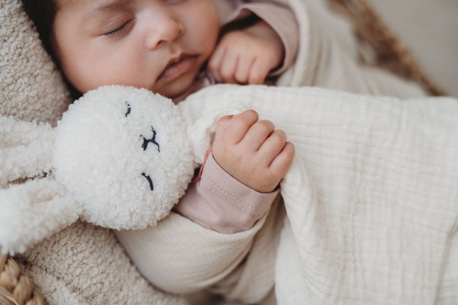 Sleeping newborn baby clutching soft white bunny lovey toy while wrapped in cream blanket.