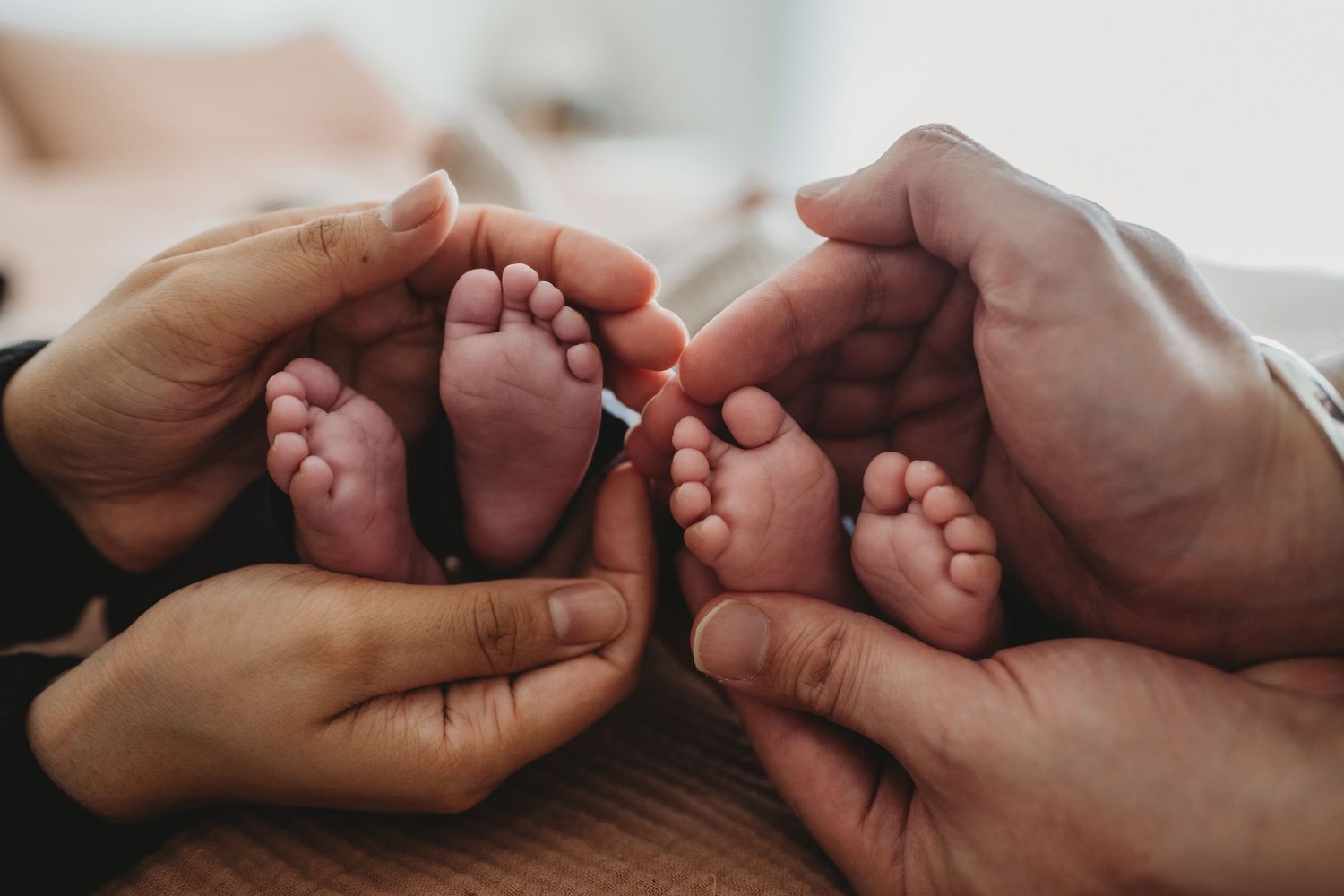 Two pairs of adult hands cradling two sets of tiny newborn twin baby feet together.