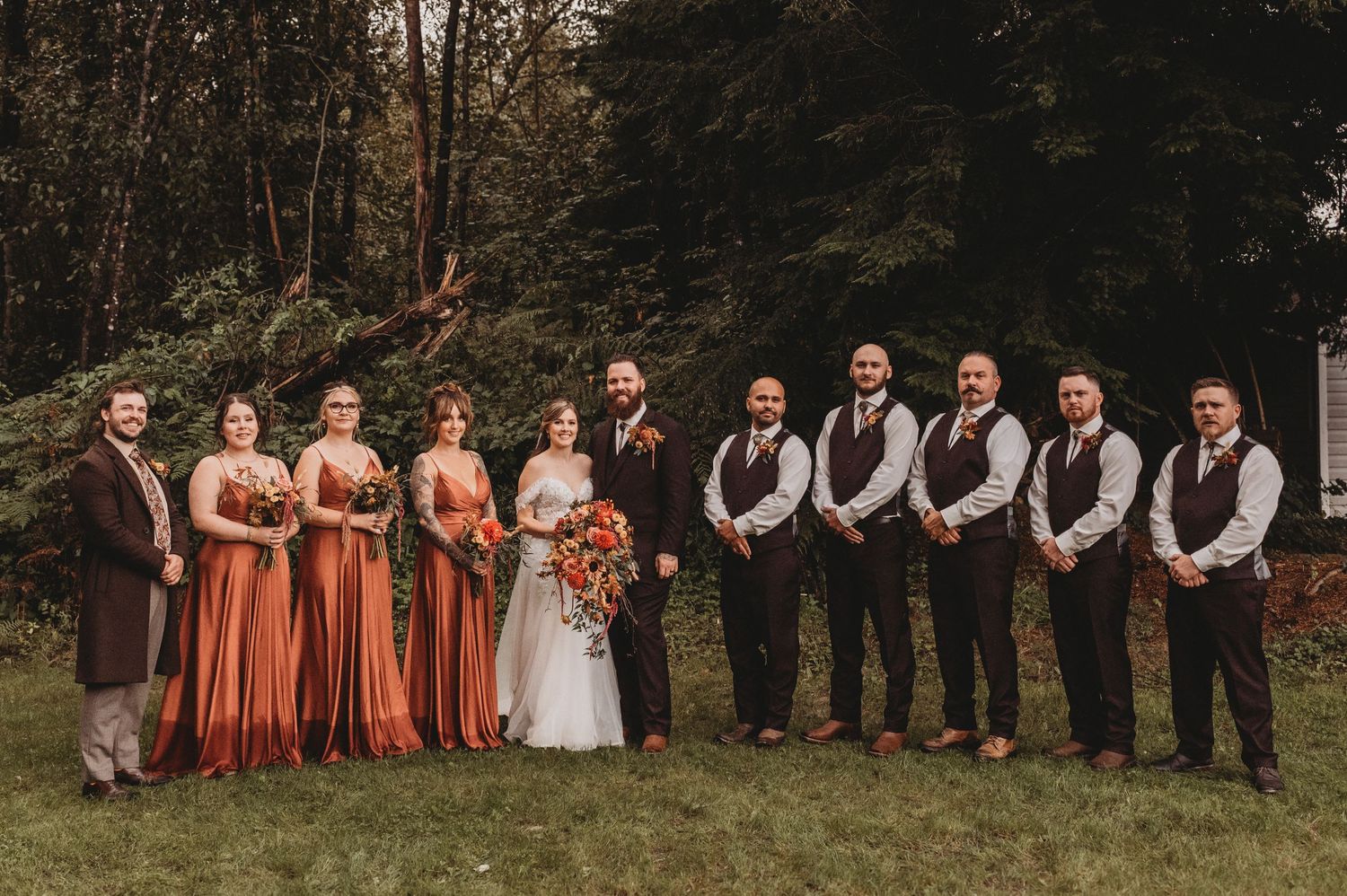 Wedding party in rust bridesmaid gowns and brown vests poses outdoors in forest setting.