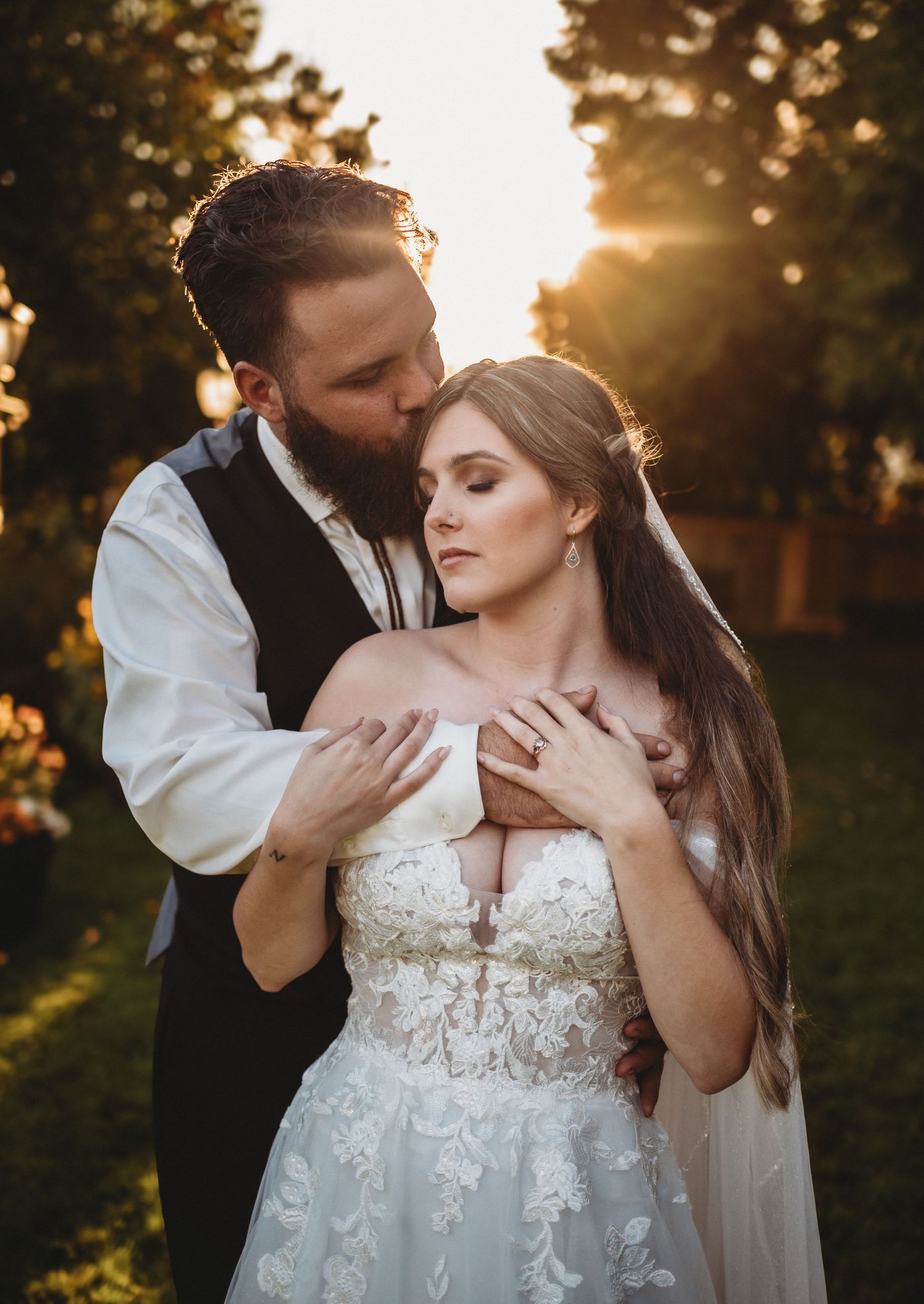 Groom kissing bride on the cheek while embracing her from behind in warm glowing sunset garden setting.