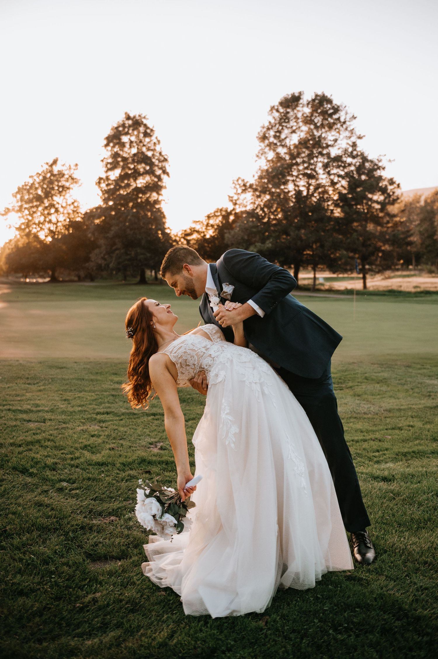 Bride and groom share a romantic dip kiss at golden hour on a lush green lawn.