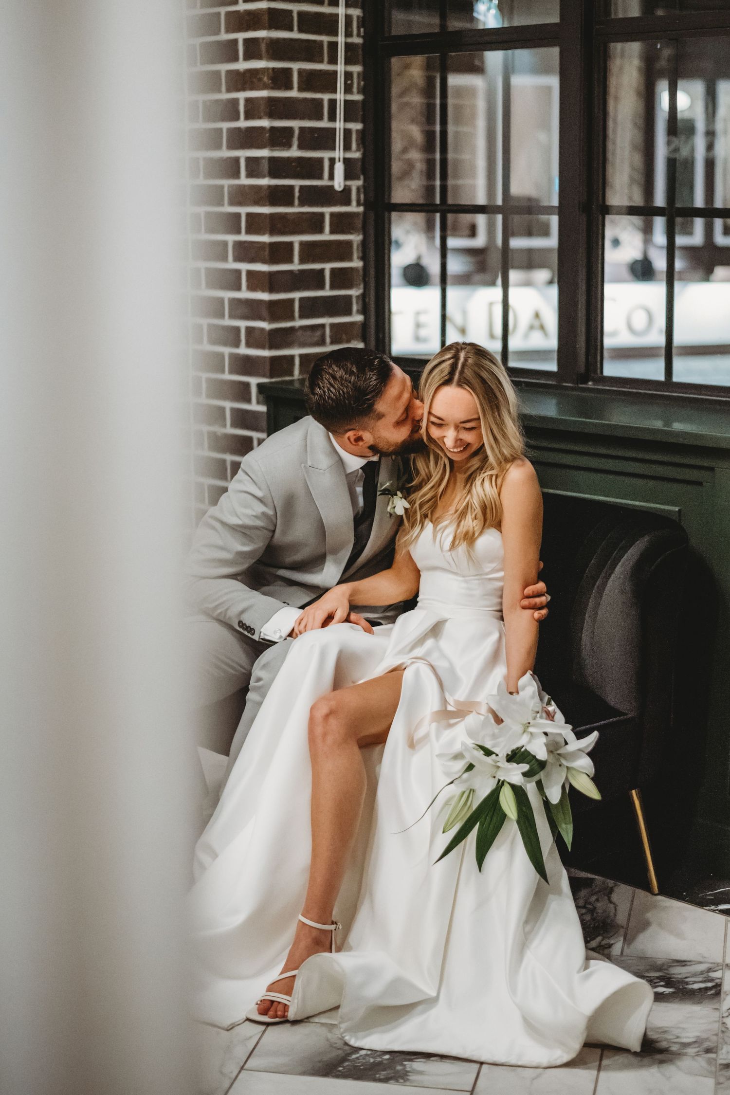 Bride and groom share a romantic kiss while seated together, bride holding a white lily bouquet in a elegant wedding setting.