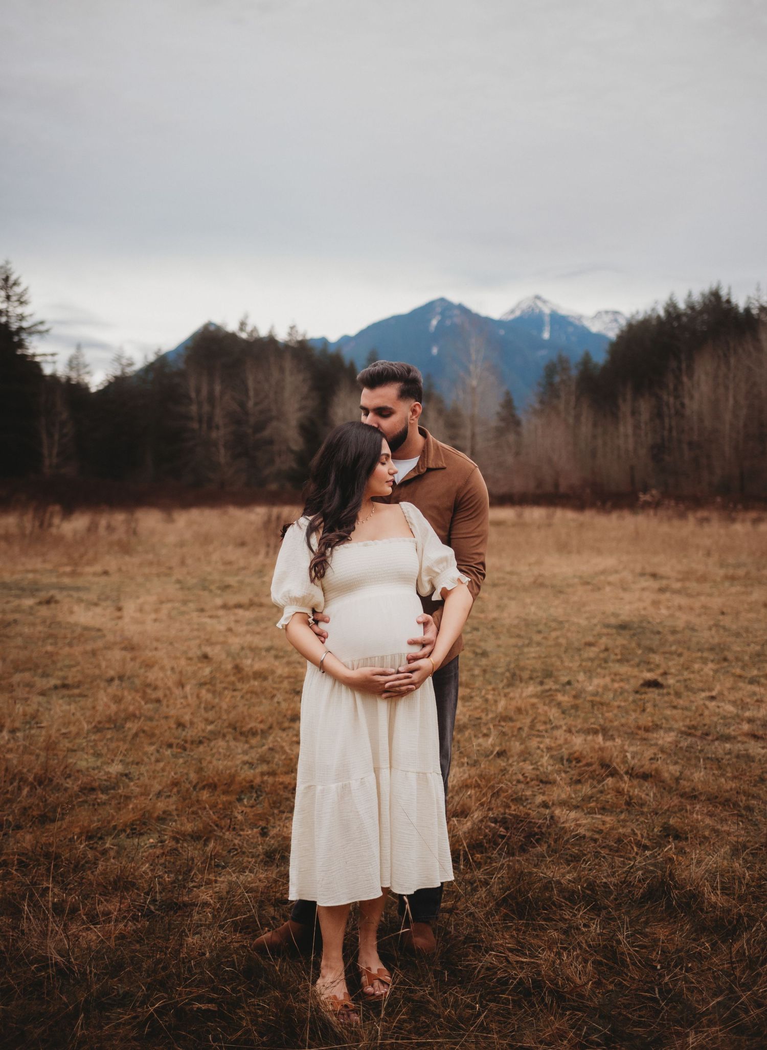 Couple maternity photo in a mountain meadow, man embracing pregnant woman in white dress outdoors.