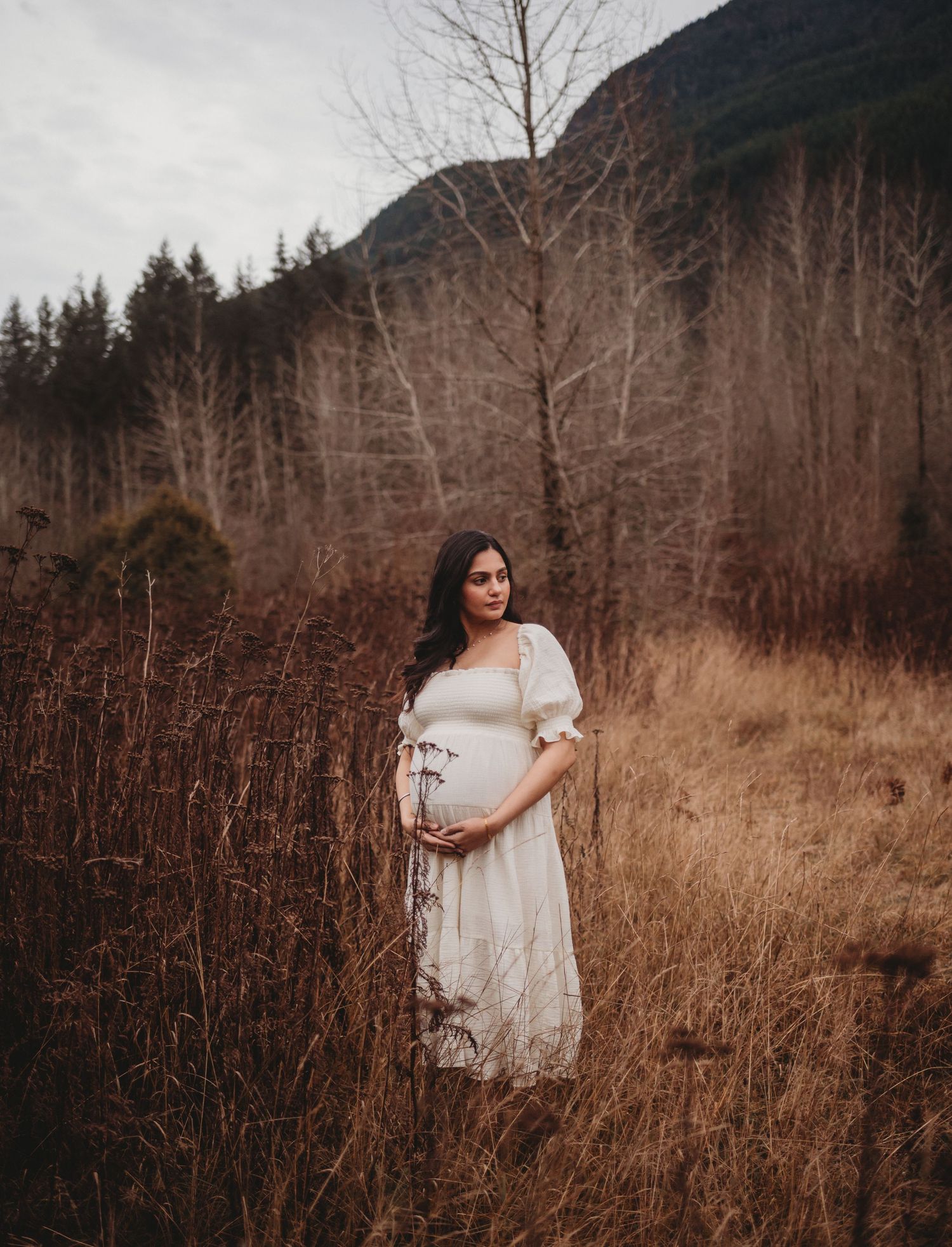 Pregnant woman in white lace dress holding belly amid tall dry grass with forested mountains behind.
