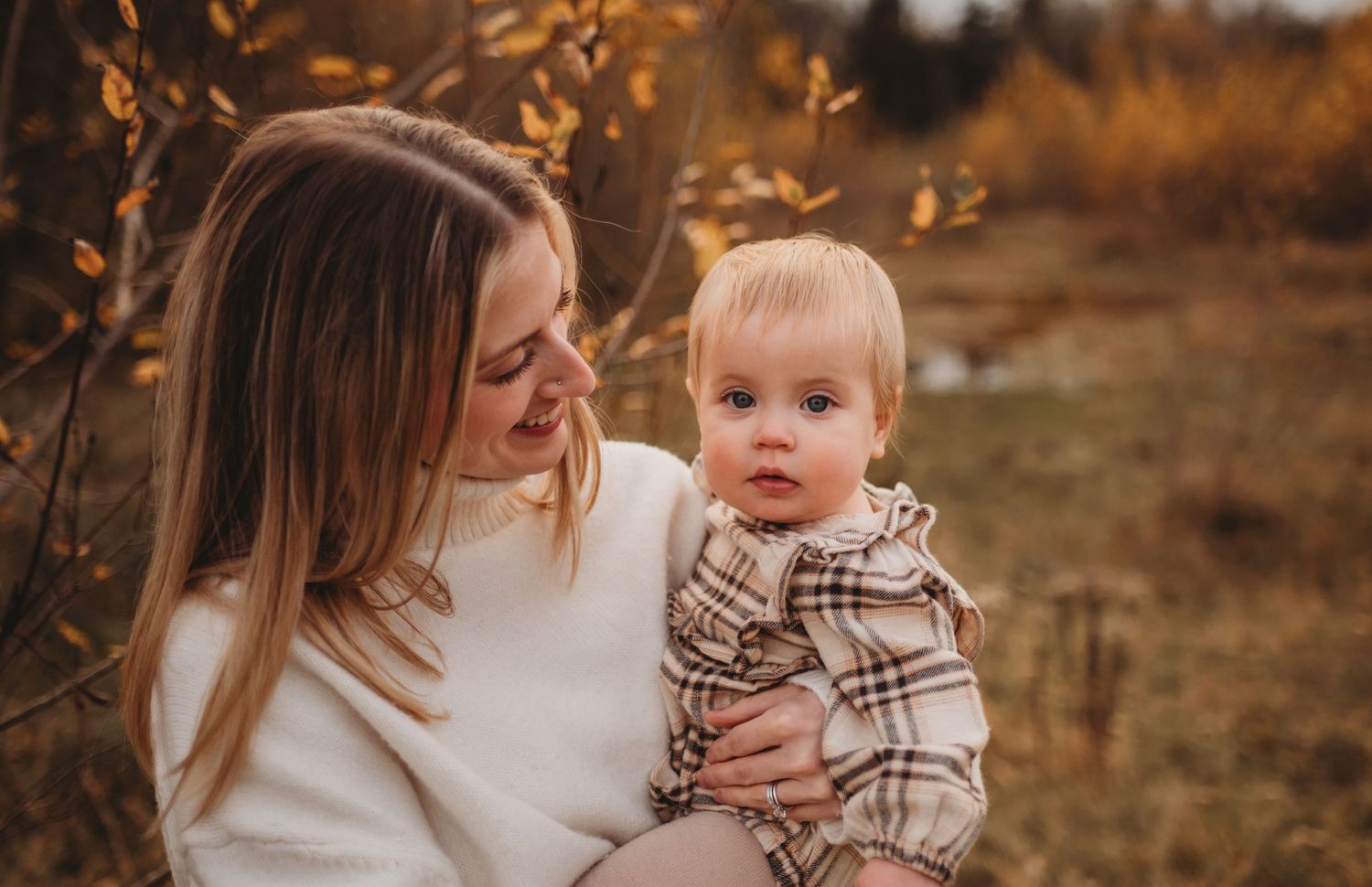 Young mother holding adorable baby in plaid outfit among golden autumn foliage during fall session.