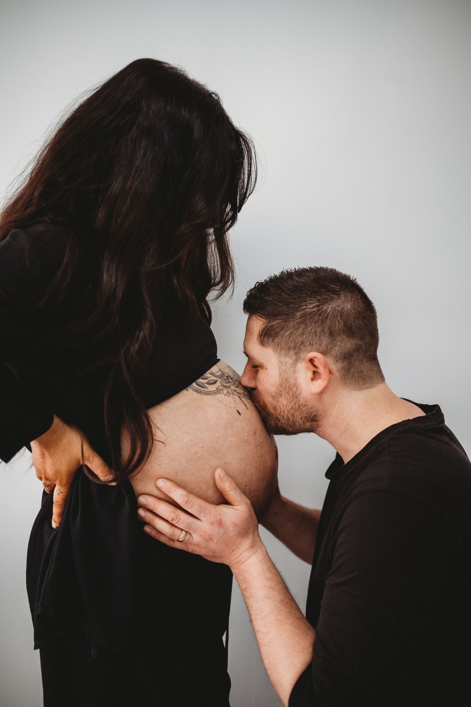 Man kissing pregnant womans tattooed baby bump during maternity photoshoot.