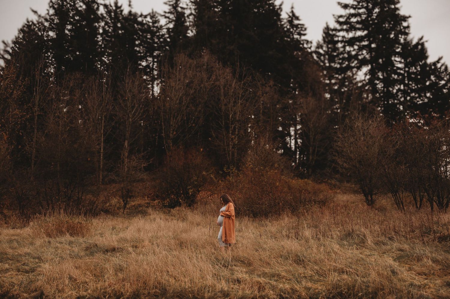 Expecting mother in orange coat standing in dry grass near forest.