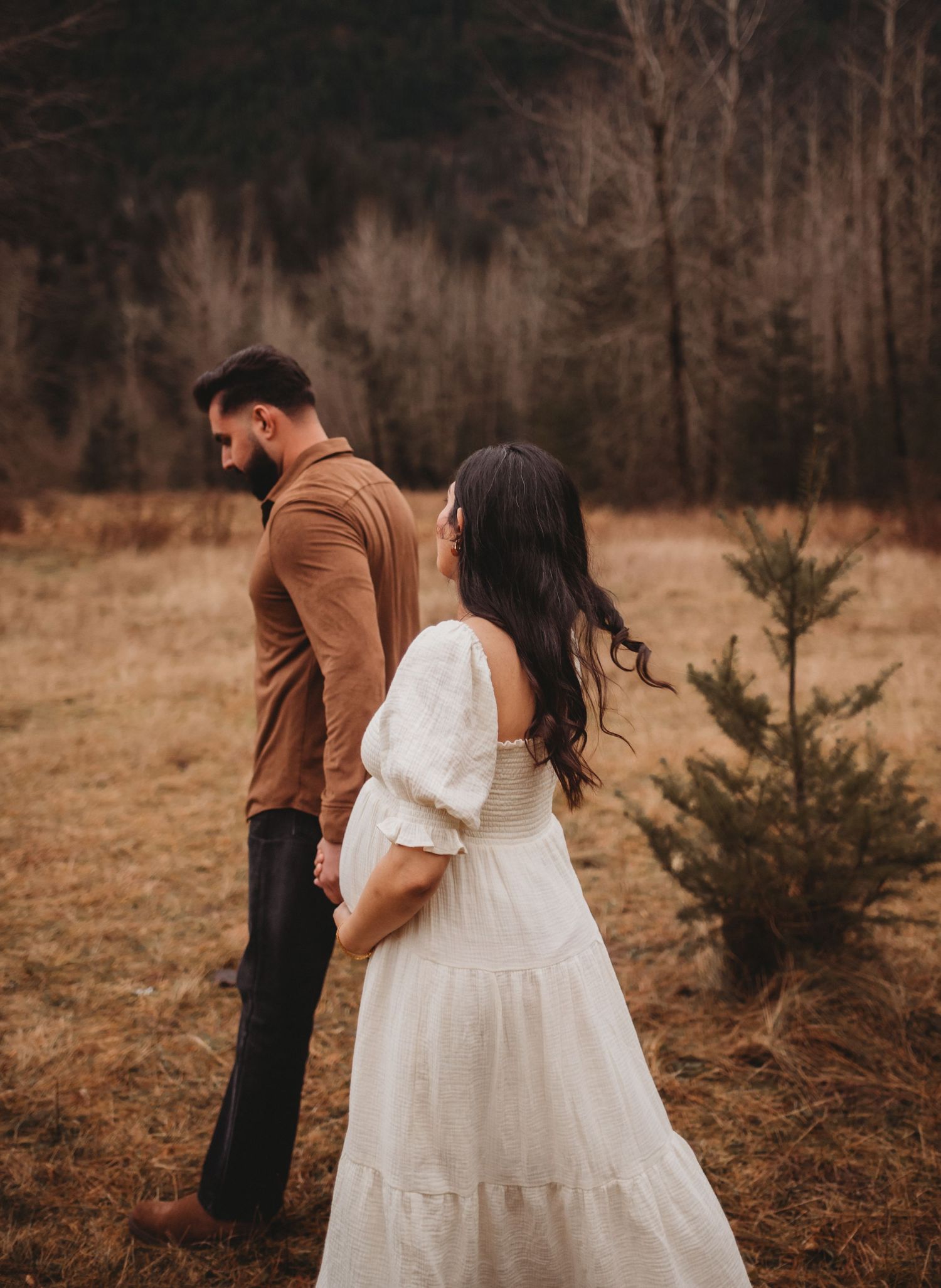 Pregnant woman in white dress holding partners hand in moody autumn forest field.