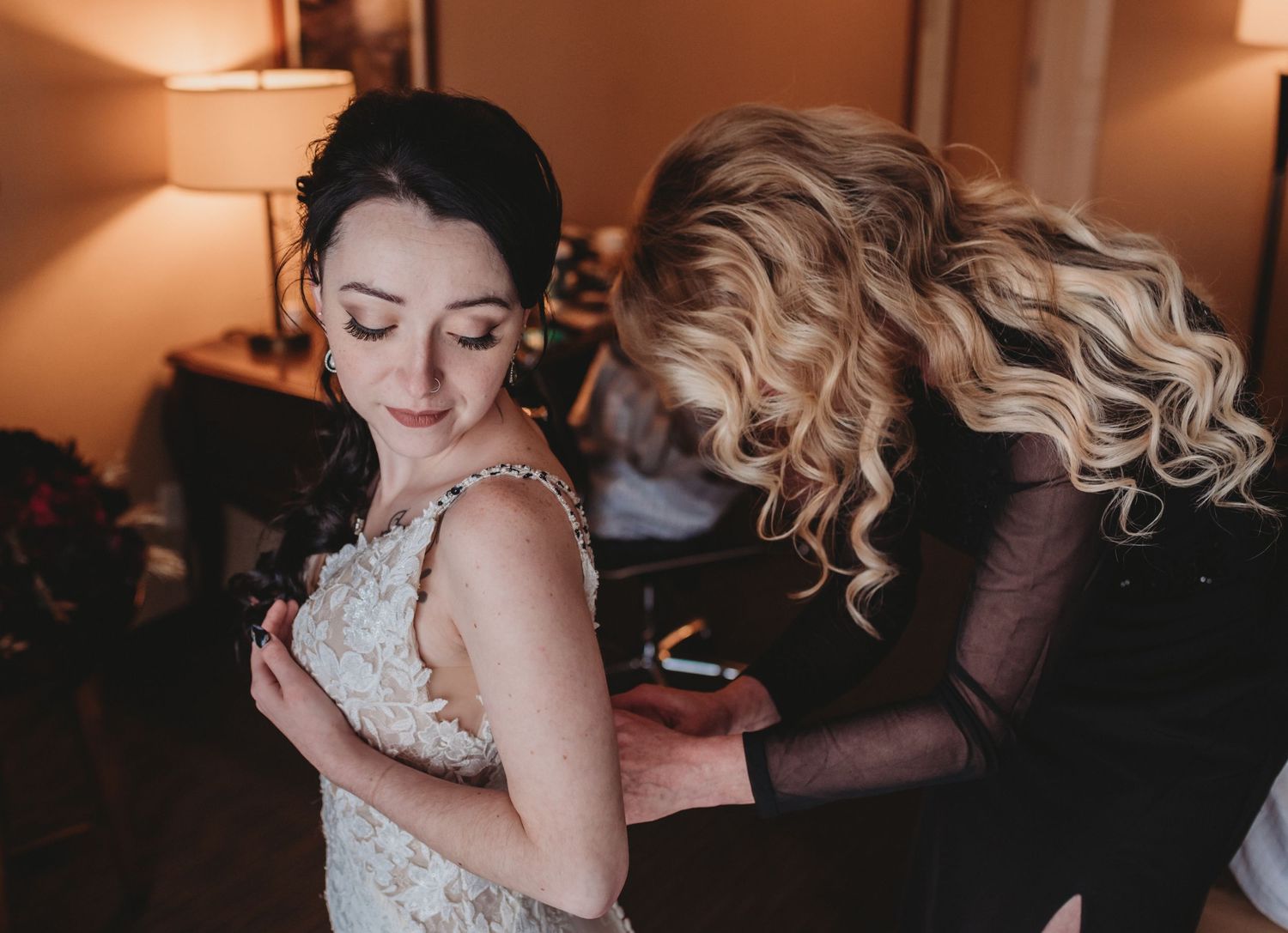 Bridesmaid helping bride fasten her lace wedding dress in a warmly lit bridal preparation room.