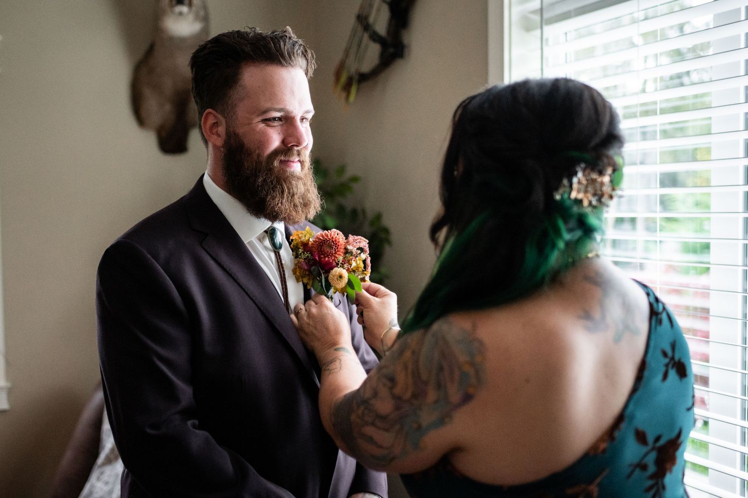 Bride with green hair and tattoos pins a colorful boutonniere on bearded groom in black suit.